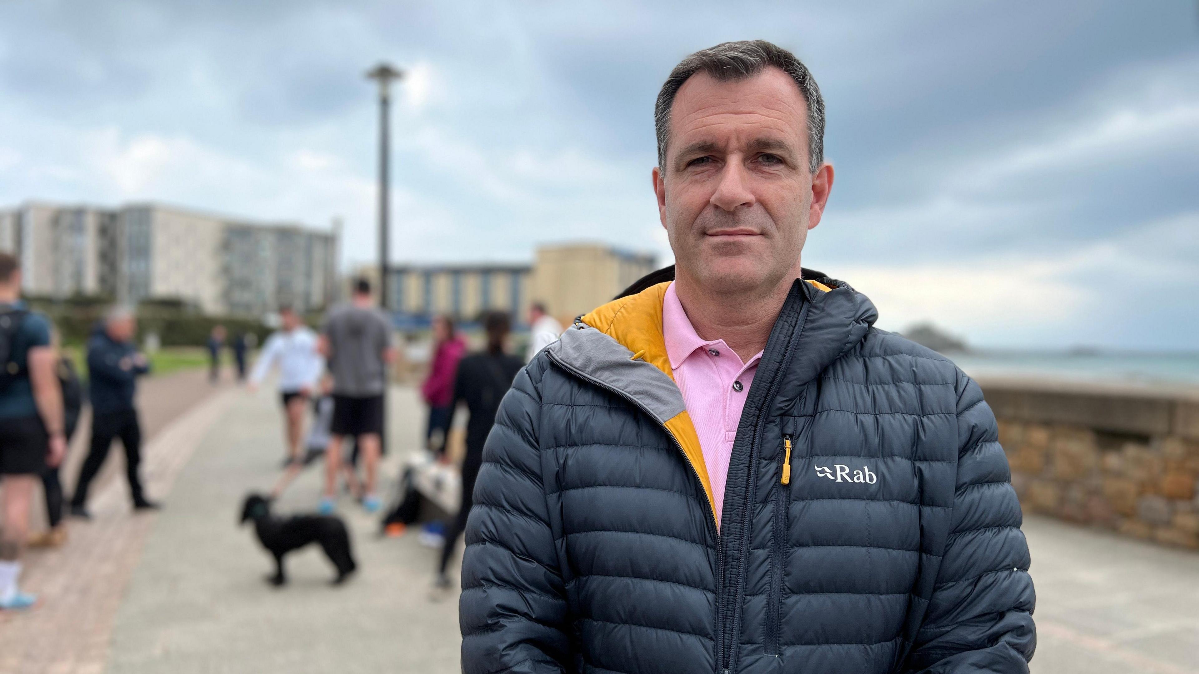 Paul looks at the camera as people sit on a stone bench behind him. He is wearing a black puffer jacket and a pink polo shirt under it.