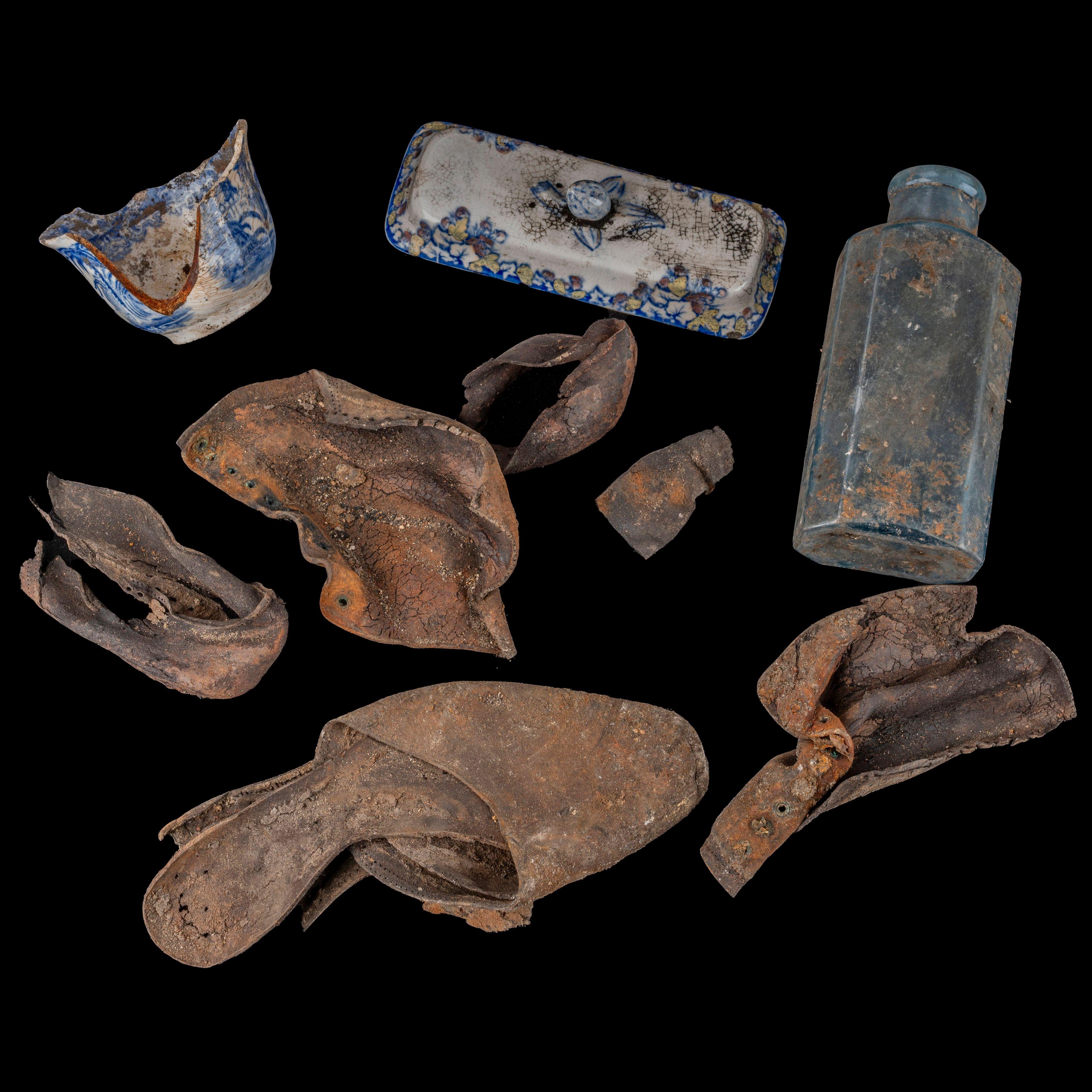 A very old looking shoe, bottle and butter dish are laid out on display against a black background.