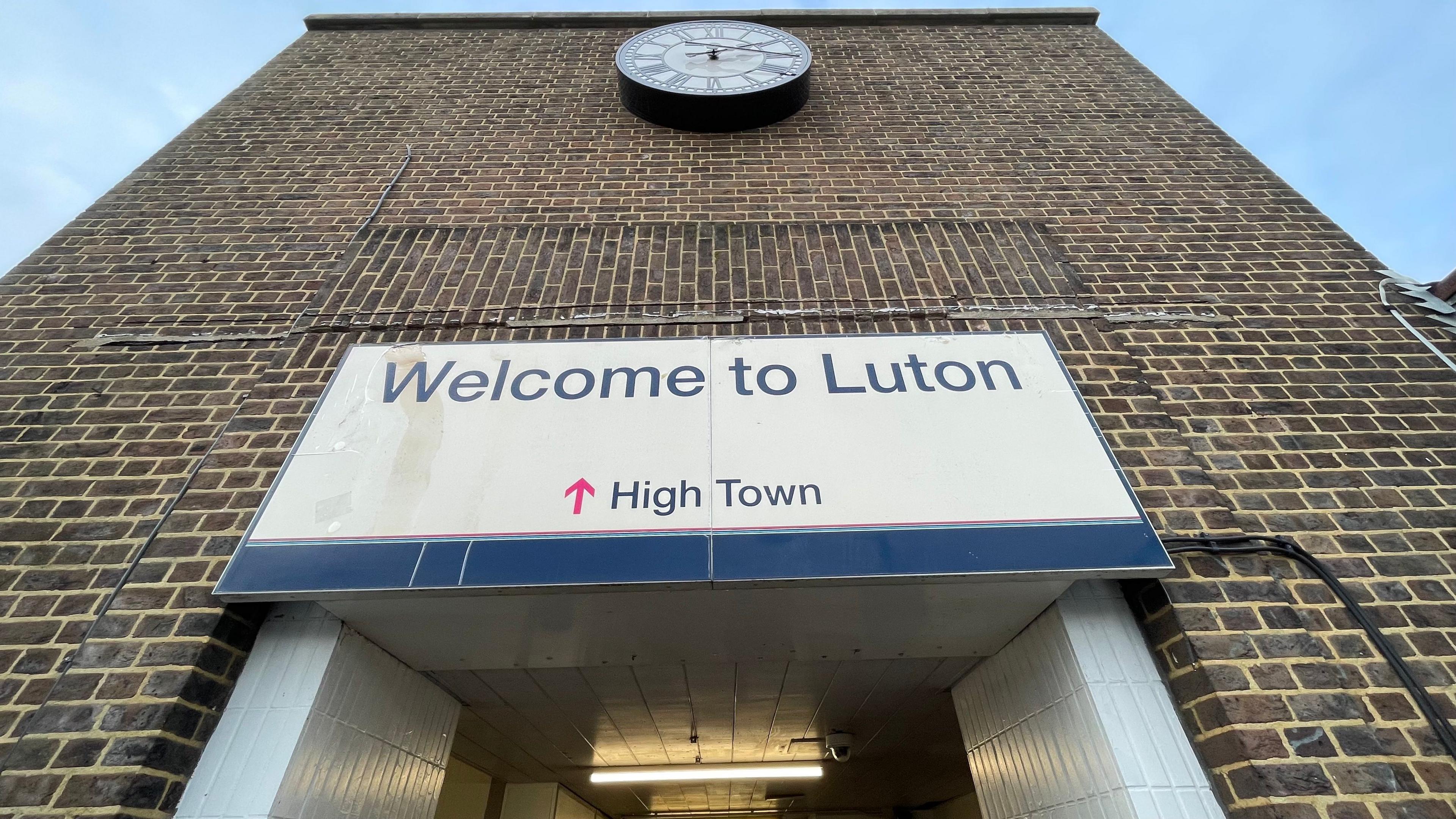 Looking up an imposing brick wall, with a "welcome to Luton" sign hanging over a doorway, which is only partially visible. Higher above the sign is a clock, with a white face and Roman numerals.