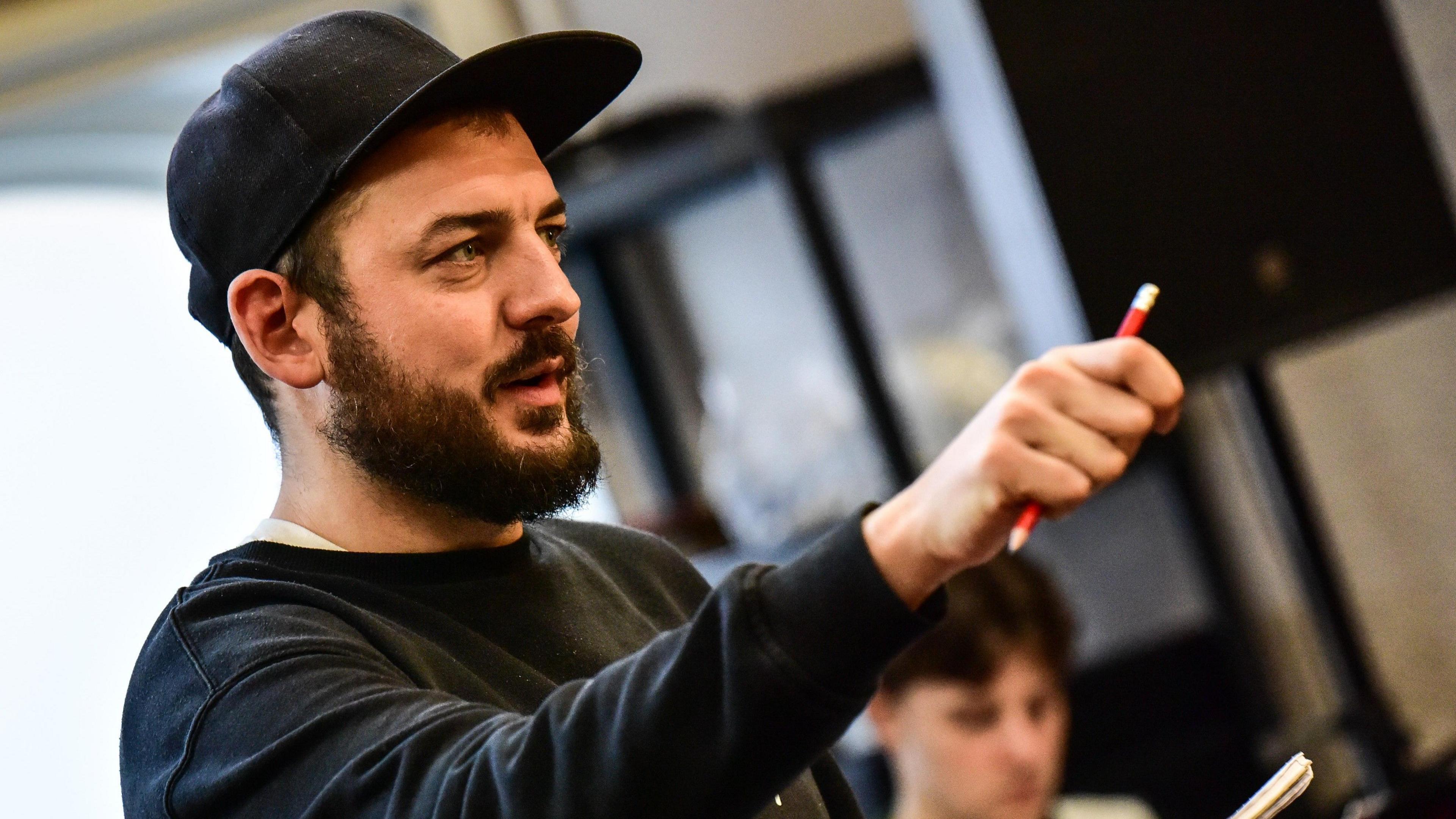 A man, with a brown beard in a black jumper and a black cap holds up his right arm holding a red pencil.