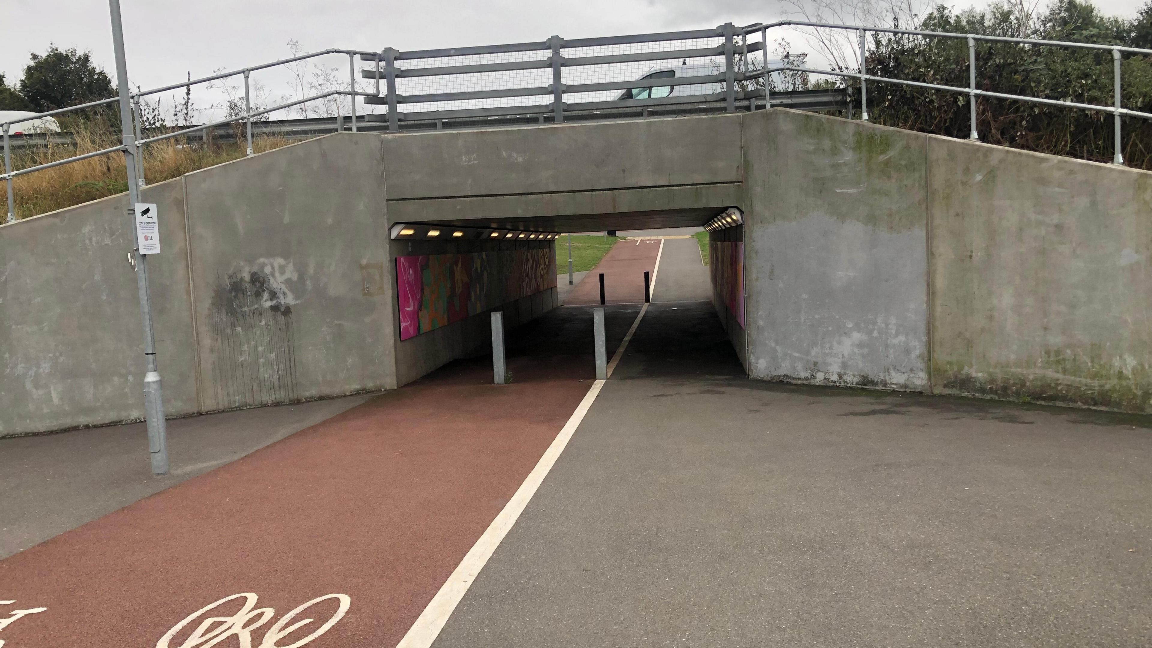 A grey concrete underpass at the Ely leisure village that goes beneath the A10 between Ely and Littleport. It is about 4-meters wide and 6-metres long and has red tarmac one side for cycles and grey the other for pedestrians. It has lights on the inside walls and art work can be seen on the walls of the subway.
