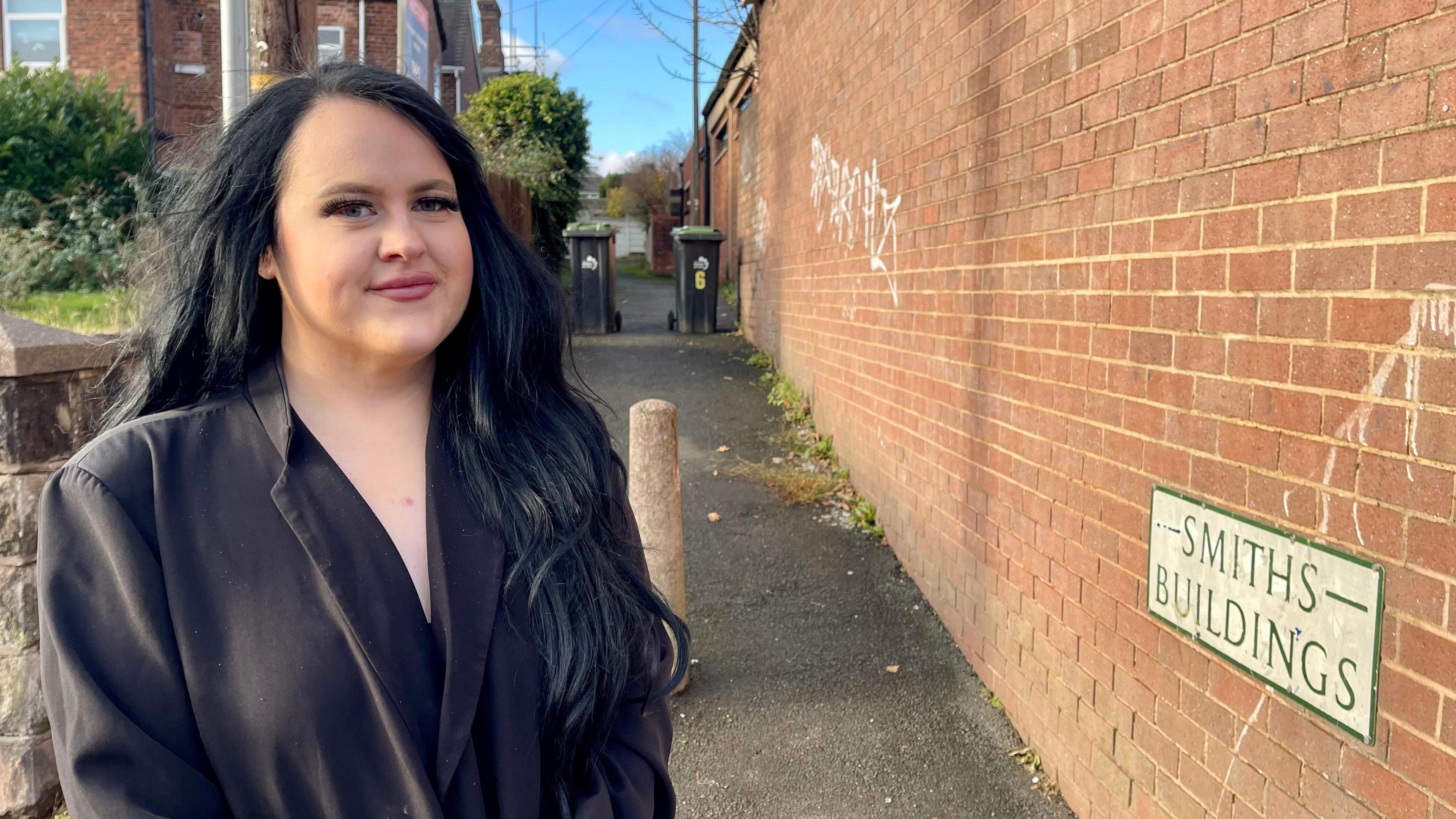 A woman with long black hair and a black blazer stands in front of an alleyway. There is a brick wall along the right one side of the alleyway and a row of houses along the other. There is a stone post in the middle of the alley and two wheelie bins further along. A street sign reads Smiths Buildings. 