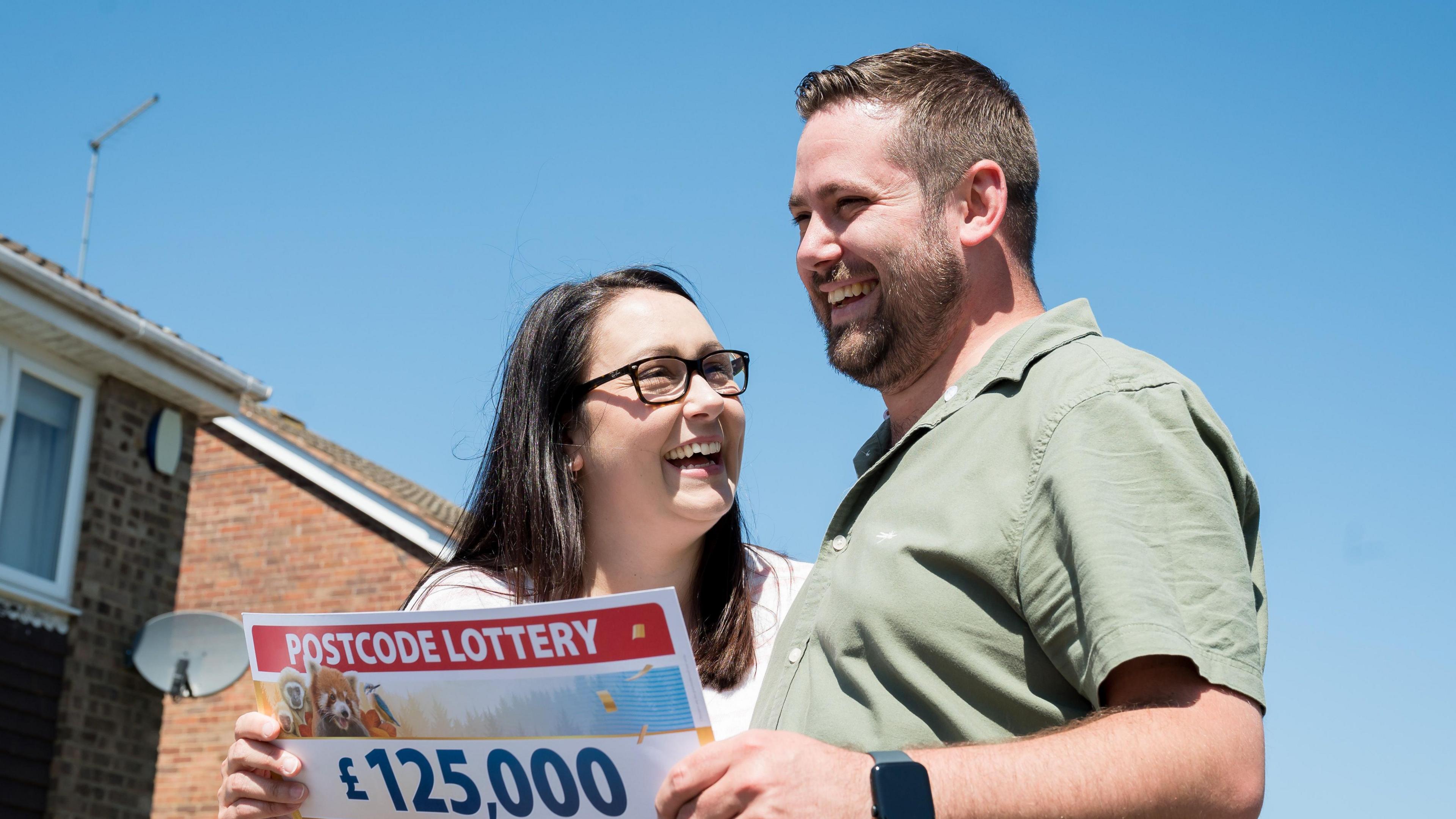 Nathan Brown with short dark hair and beard wearing a green shirt and holding a cheque for £125,000 which is also being held by Jade Dunkley who has long dark hair and glasses and is wearing a white top. There is a modern brick house behind them.