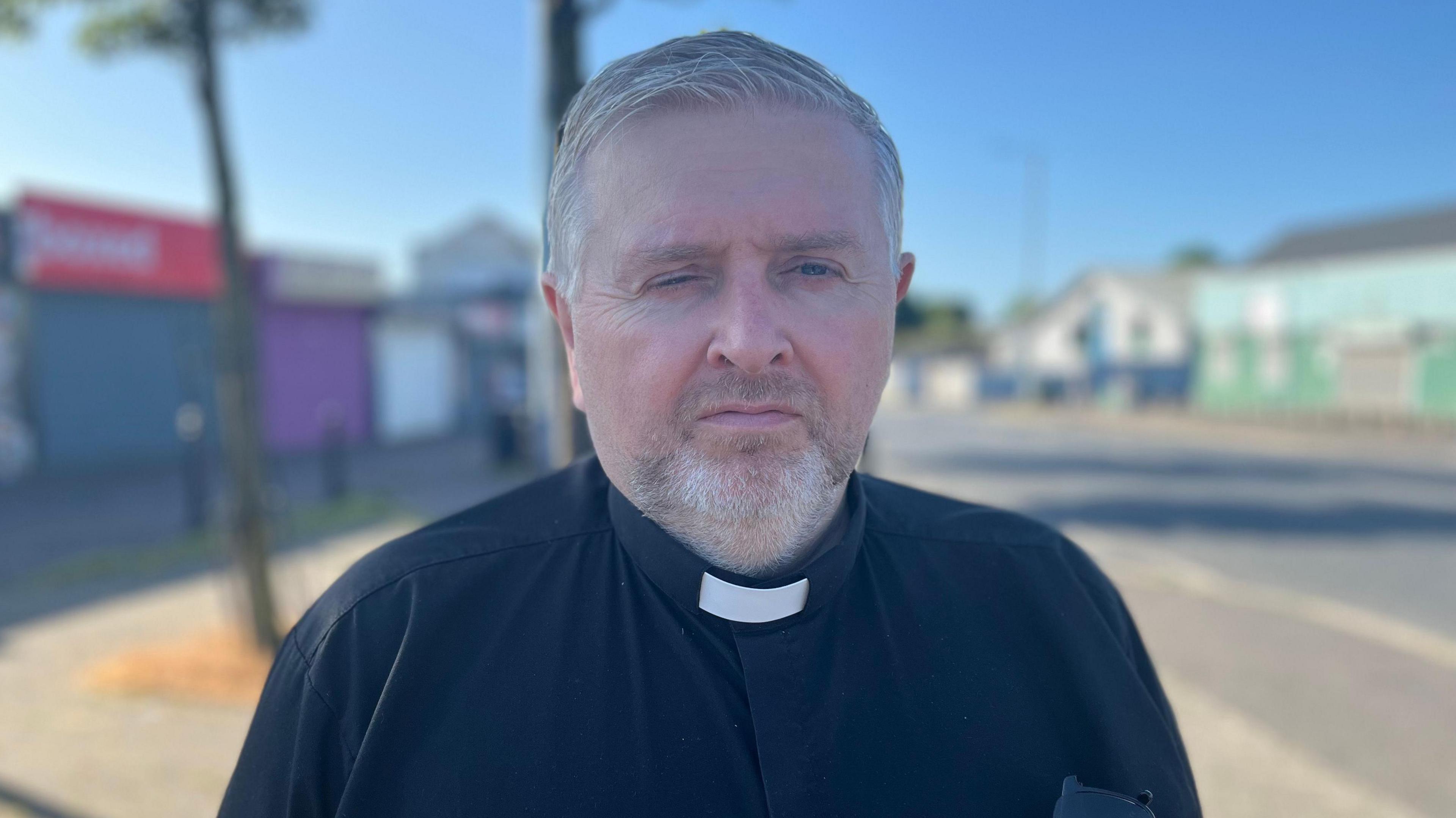 Fr Gary Donegan is standing in front of a sunny row of shops. He has grey hair and a grey beard, and is wearing his black priest attire, with white collar.