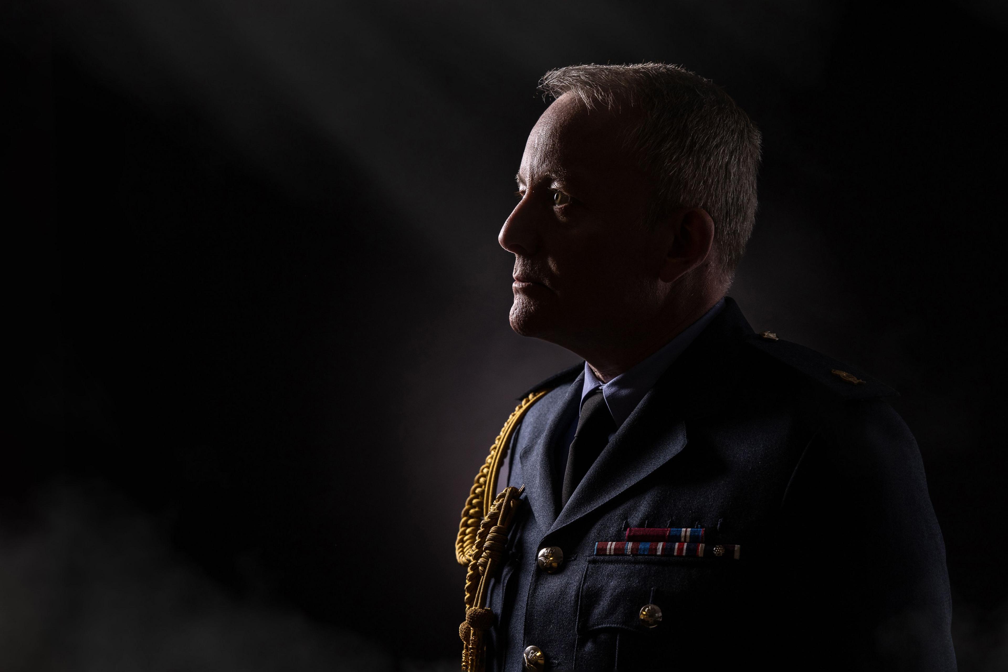 A mostly dark photo, with the Air Chief Marshall in profile to the right of the picture.  The decoration and medals on his jacket are the only colour in the photo and catch the light.  Sir Richard Knighton's face in profile catches the light on the very front of his face.