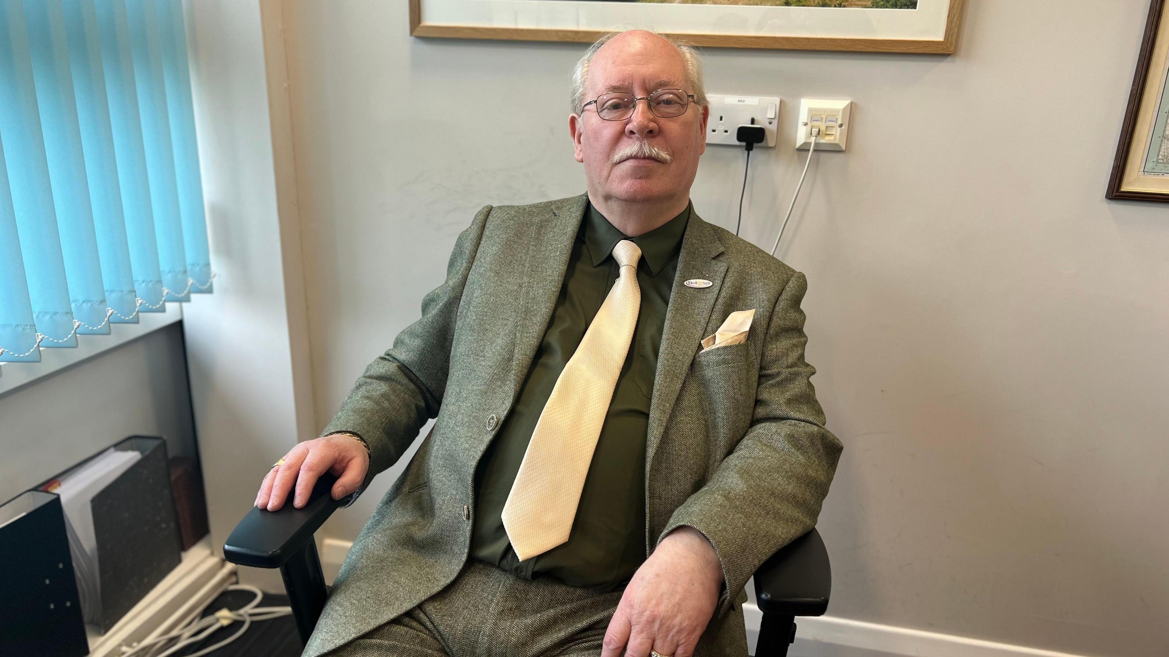 An elderly man with glasses in a light green suit and yellow tie sits at his desk in his office