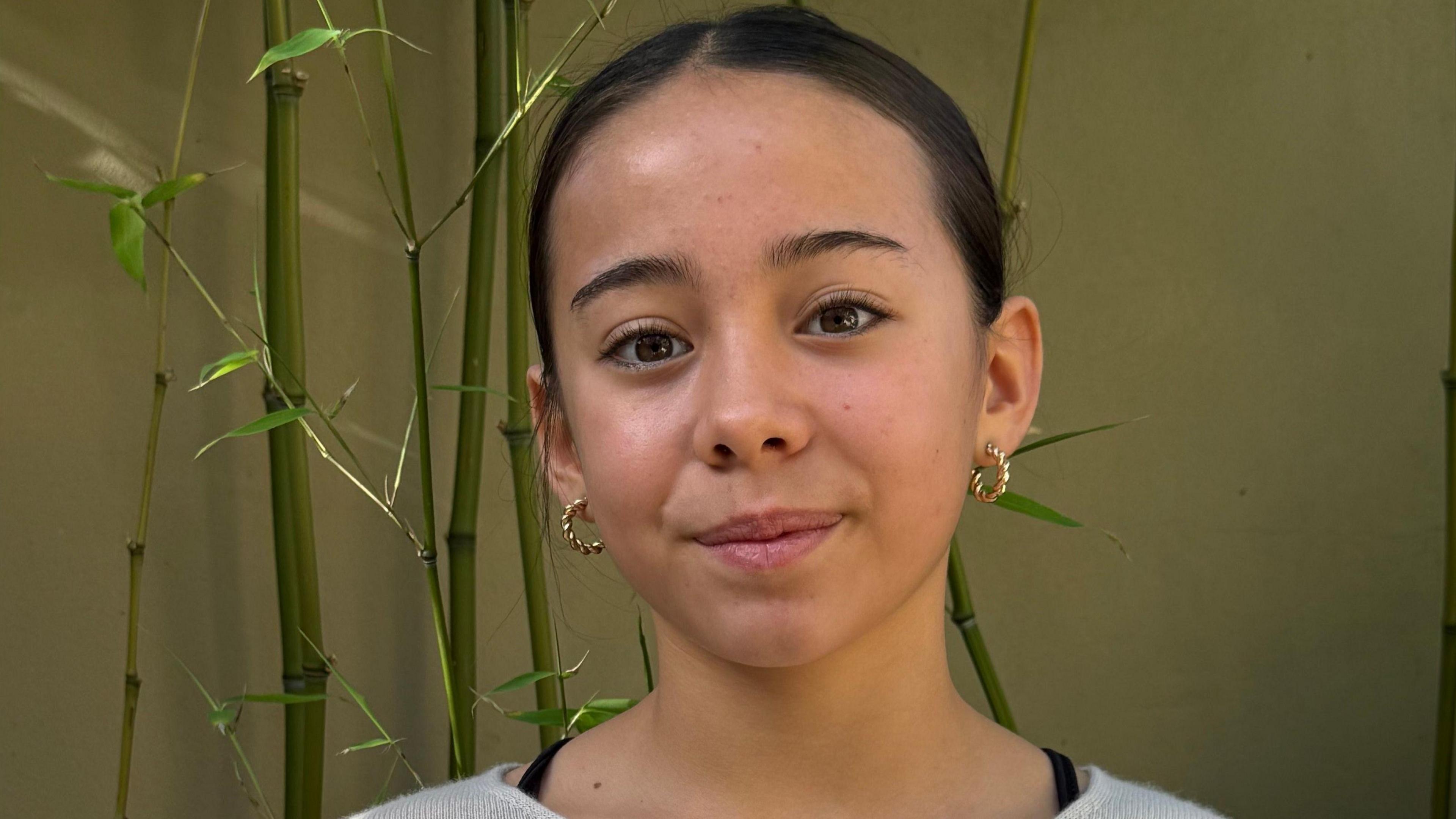 Juliette Broad is smiling at the camera. She has brown eyes and dark brown hair which is tied back. She is wearing gold hoop earrings. There is bamboo behind her.