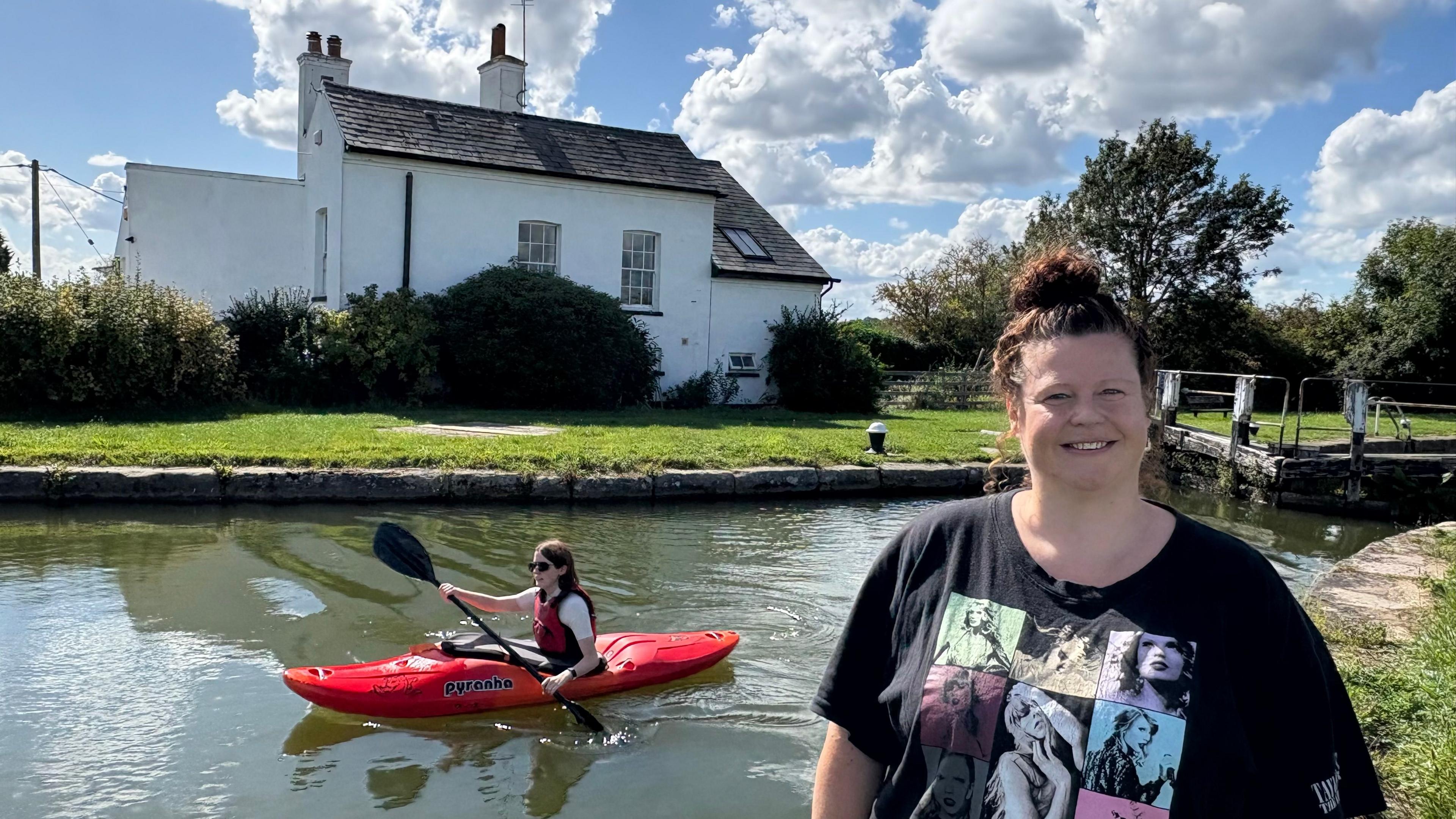 Kristina Hedley with long brown hair wearing a black T-shirt standing on the towpath beside a canal, on which a red canoe is being paddled. A two storey which house is visible on the other side of the canal with two chimneys. There is a hedge in front of the house and several clouds are visible in the sky.