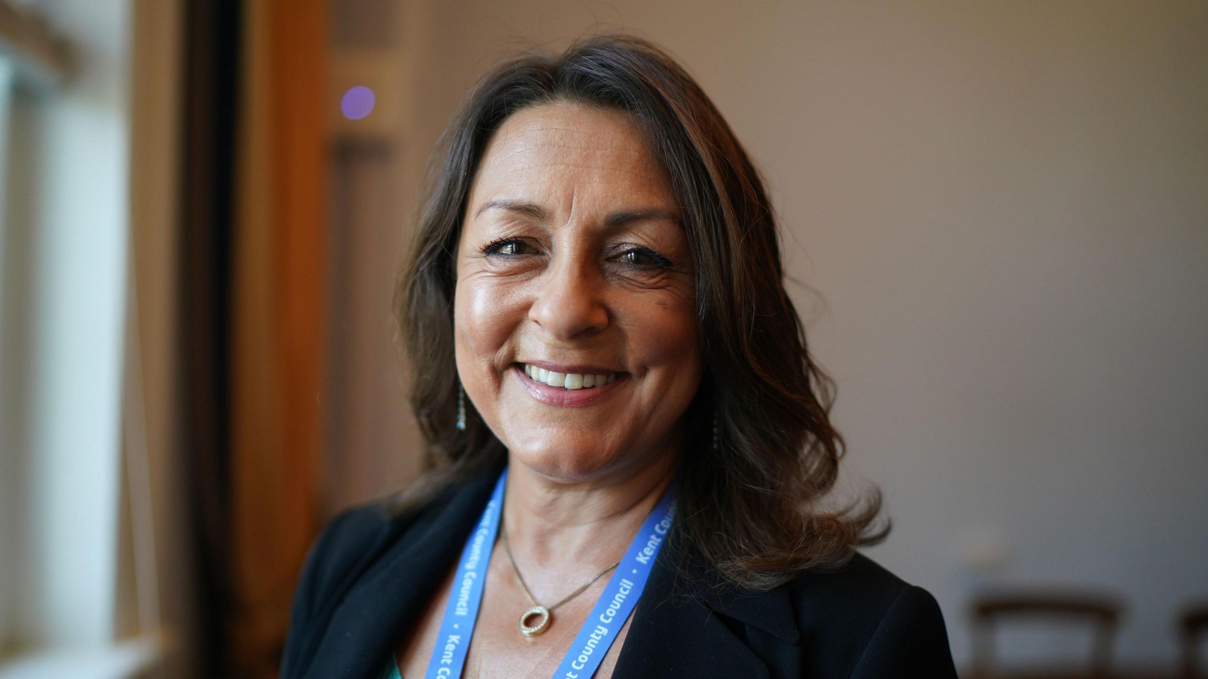 Linden Kemkaran, wearing a black jacket and a light blue Kent County Council lanyard, smiles into the camera.