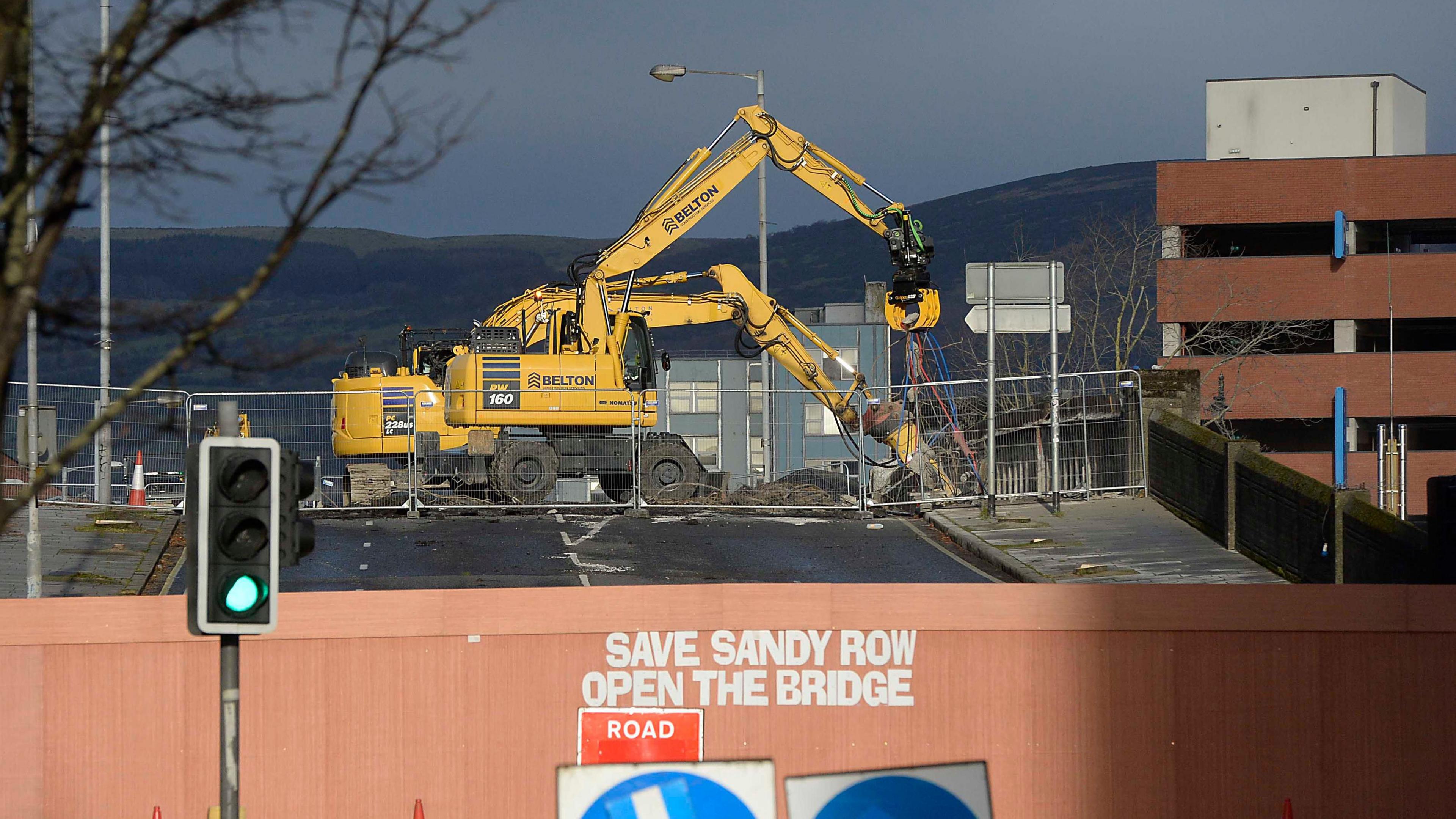 A wide shot of two yellow diggers on the Boyne Bridge in Belfast. In the foreground on the construction's wall in white letters says "Save Sandy Row, Open The Bridge".