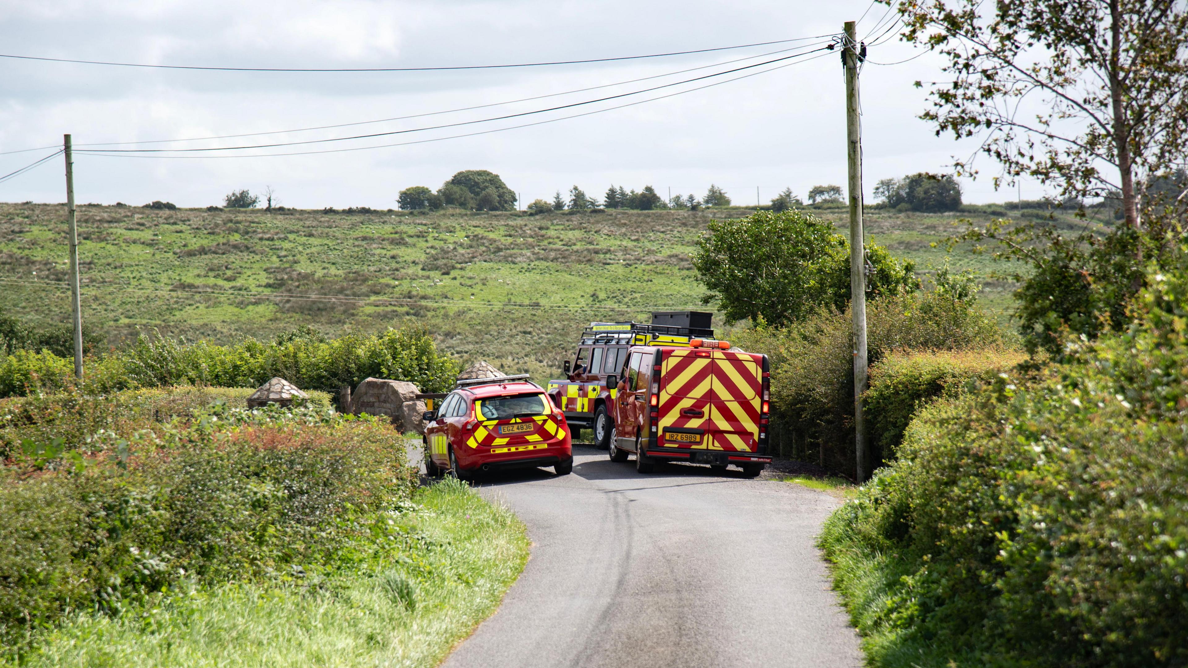 Red and yellow striped emergency vehicles are parked on a country lane which is surrounded by green fields.  