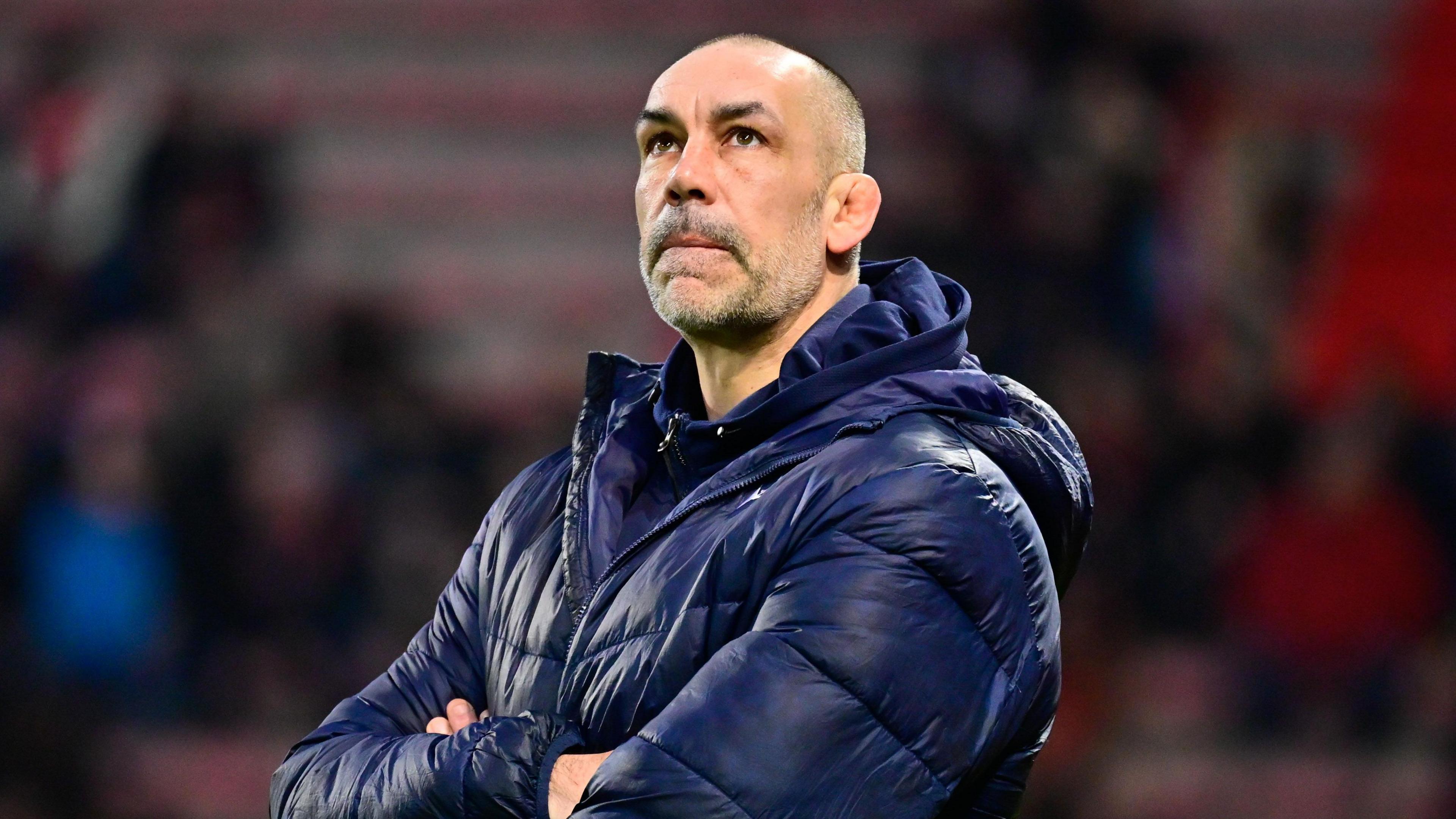Alex Sanderson standing on a pitch with his arms folded across his chest looking up towards the sky