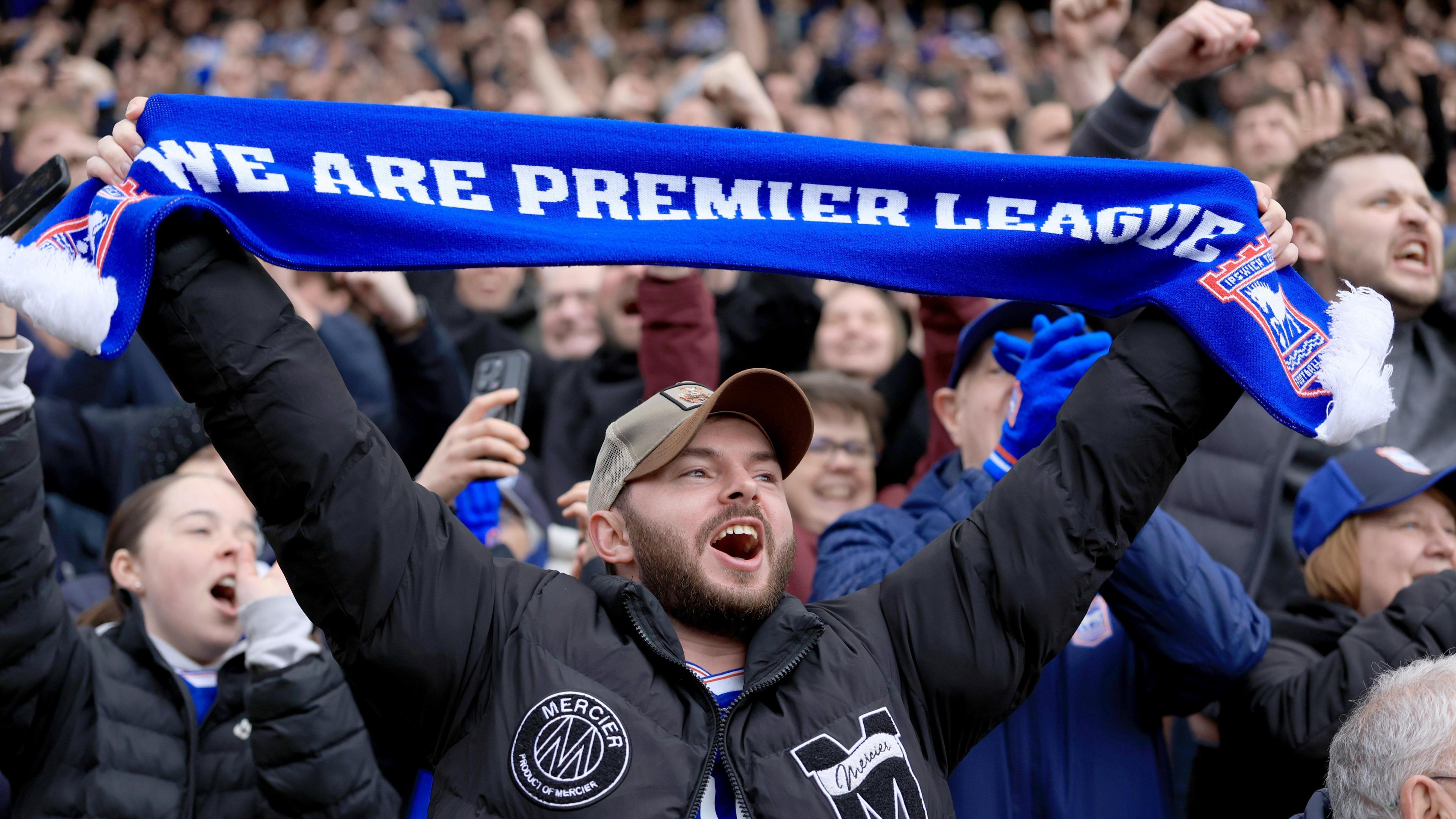 An Ipswich fan holds up a blue scarf with white writing which reads "we are Premier League" and has the Ipswich Town badge on each end. Ipswich fans can be seen cheering and chanting behind him.