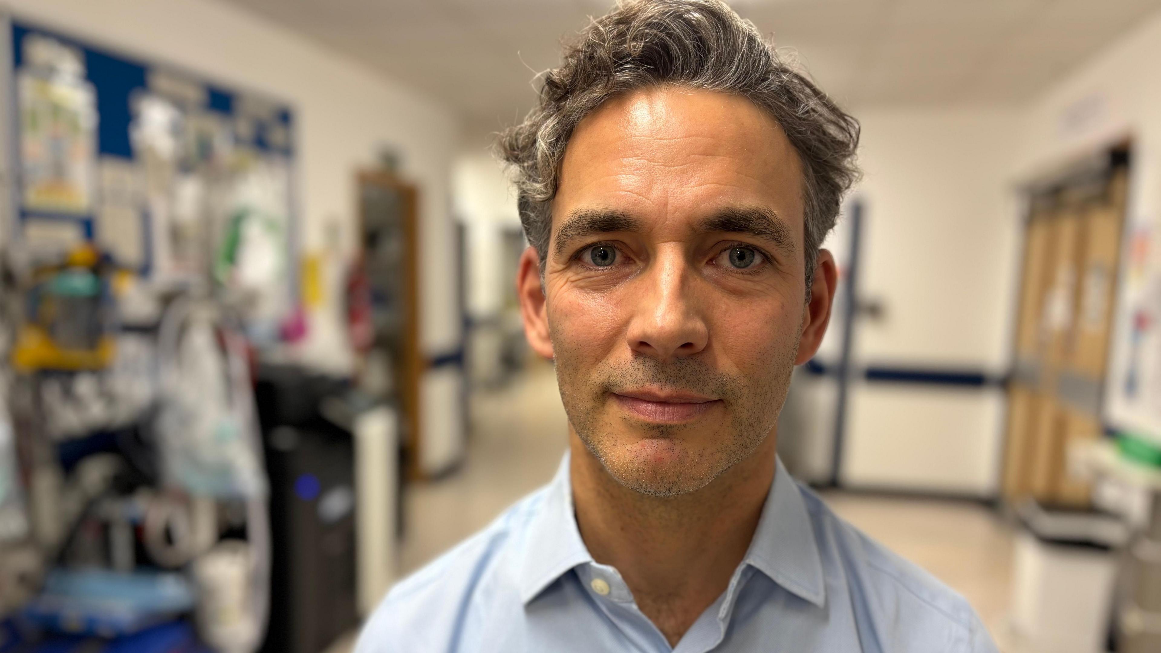 A man with short hair is wearing a light blue shirt and standing in a hospital corridor.