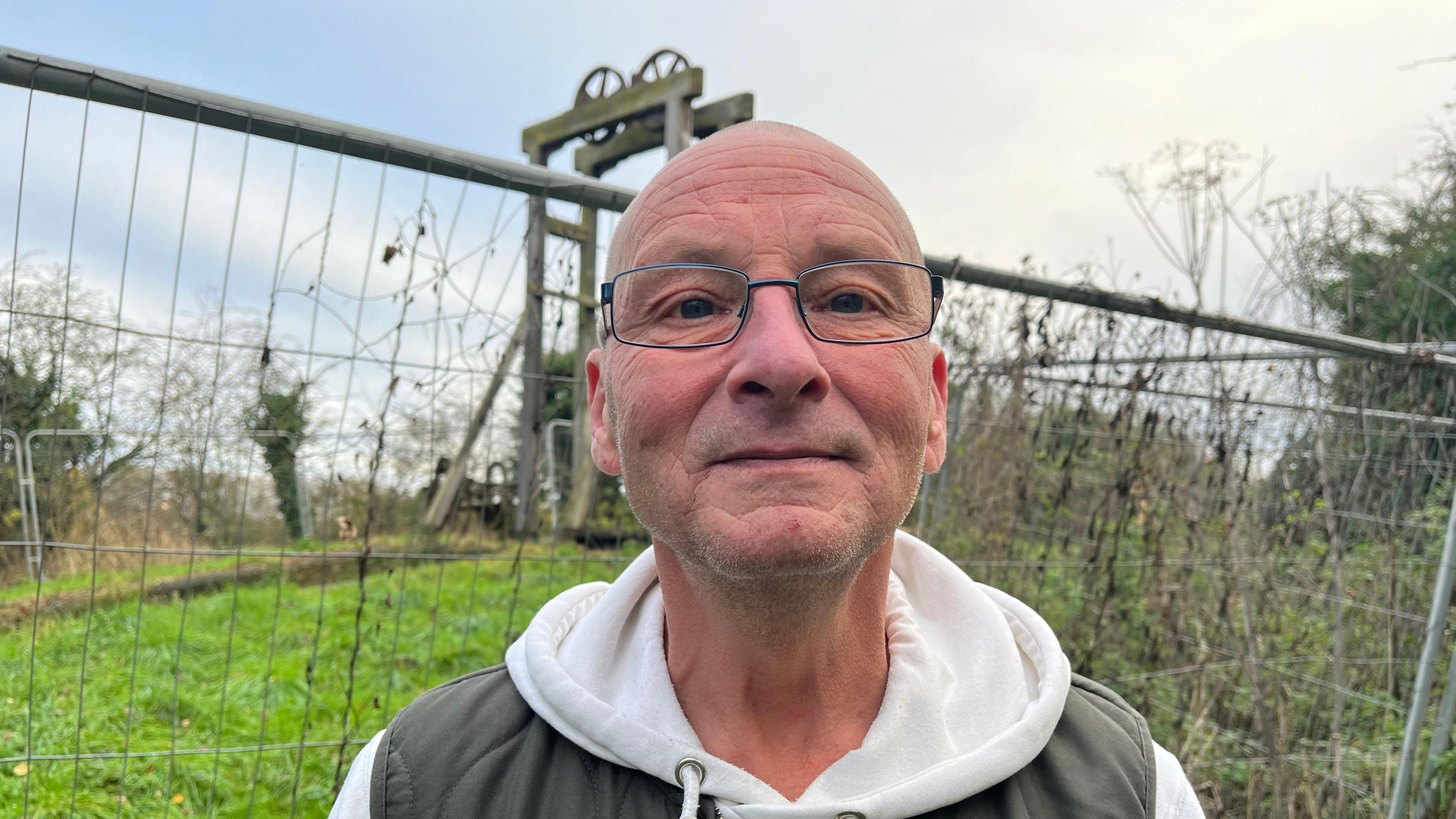 Colin looking down the camera.  He is wearing a white hoodie and a khaki gilet. Behind him is metal fencing, and beyond that you can see the Hadley Park Lock, a Grade II listed structure. The grass behind Colin is bright green and there's shrubbery over his left shoulder. The sky is blue with some clouds. 