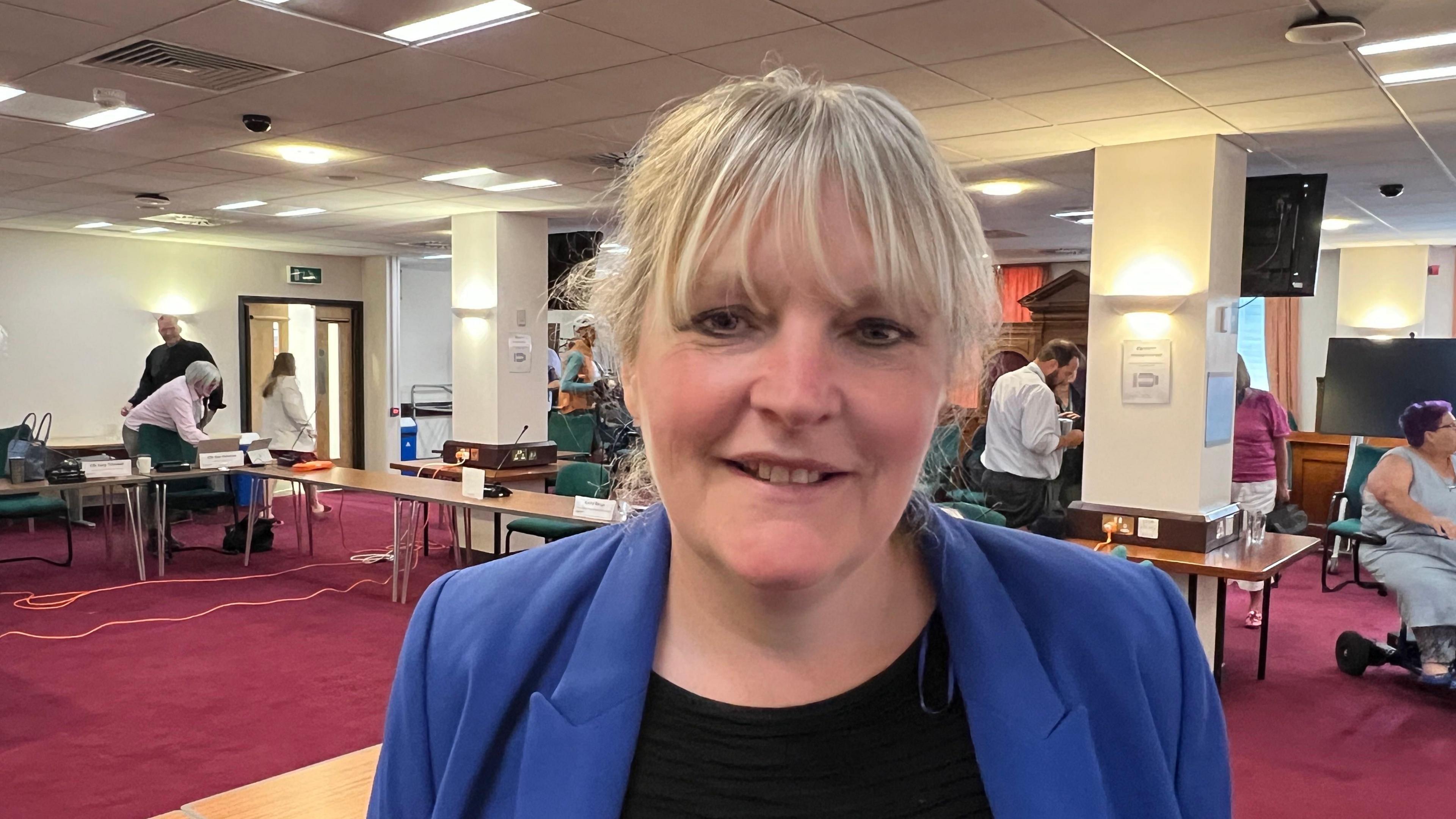 Councillor Claire Sully standing in a council meeting hall which has red carpets and wooden foldaway tables. She has blonde hair styled into a fringe, is wearing a black top underneath a bright blue blazer, and smiling at the camera.