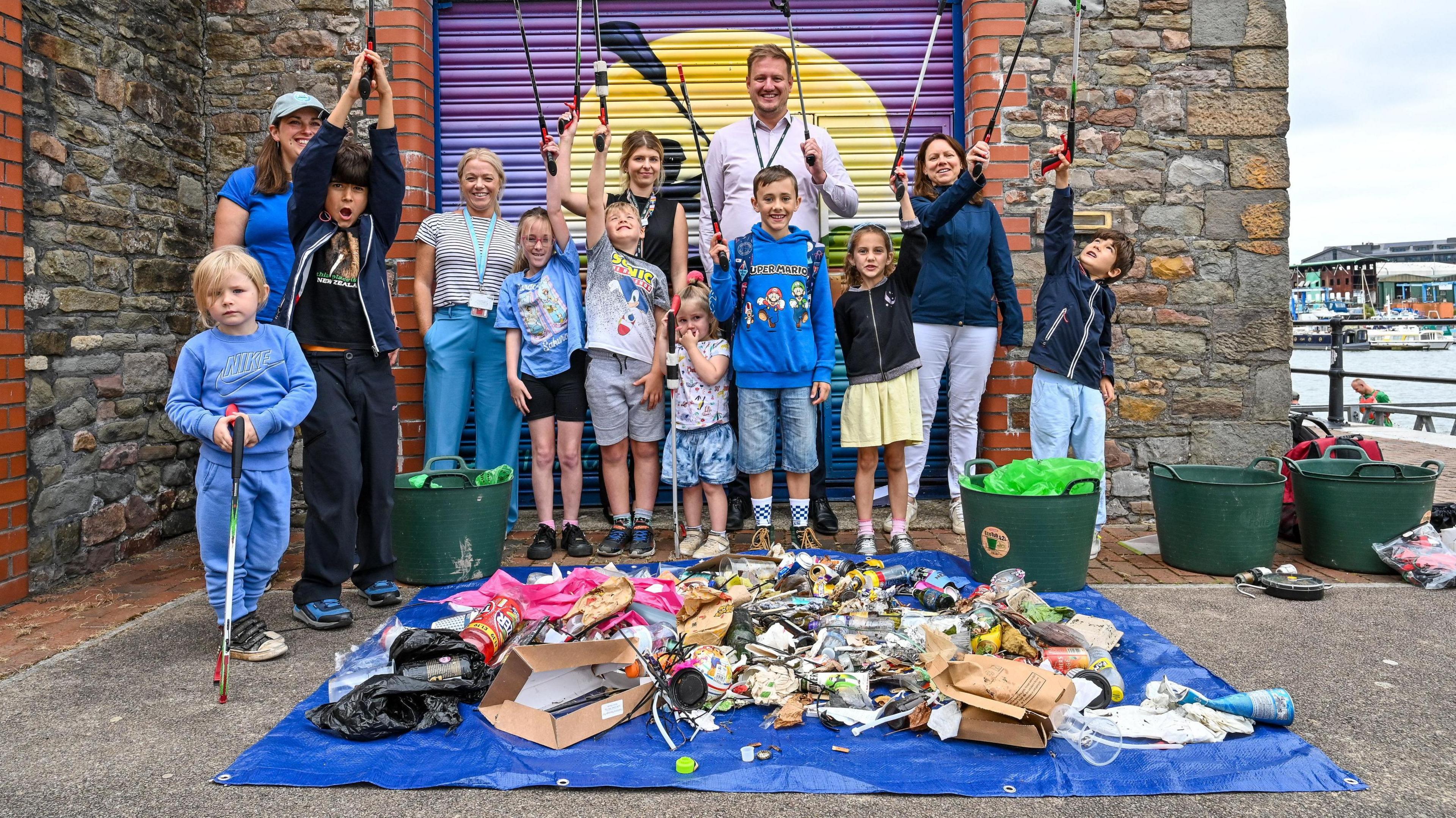 A group of five adults and eight children pose in front of a blue tarpaulin covered in trash