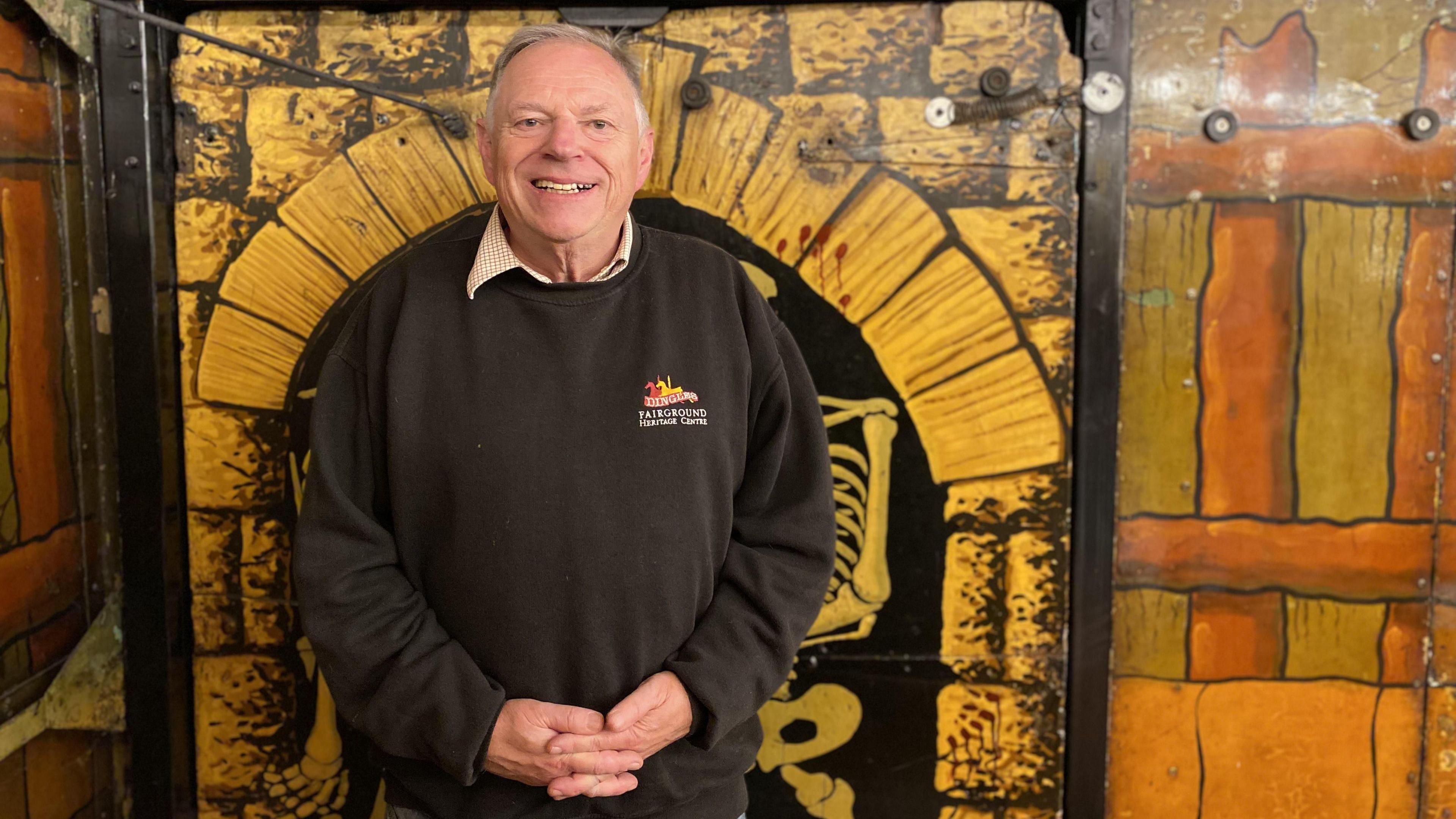 Guy Belshaw is smiling at the camera with his hands clasped in front of him. He has grey hair and is wearing a branded black sweatshirt. He is standing in front of a wall that has been painted in a cartoon style to show an arch made of stone and part of a skeleton standing under it and a wooden fence.