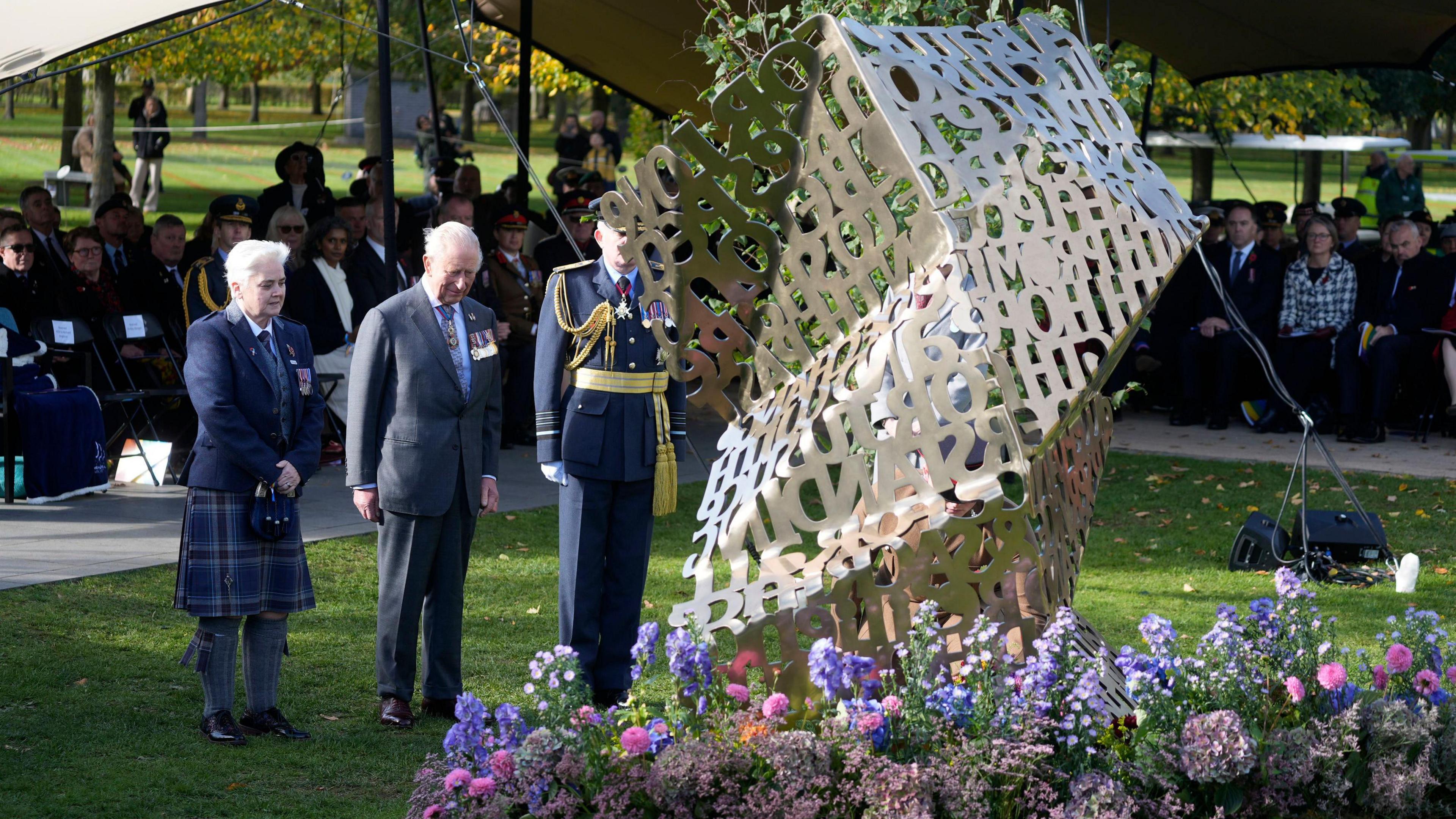 King Charles III stands in front of a bronze memorial at the National Memorial Arboretum during a dedication ceremony. Two military personnel are stood either side of the king with medals on their chests. A crowd of people is gathered on seats behind the trio.