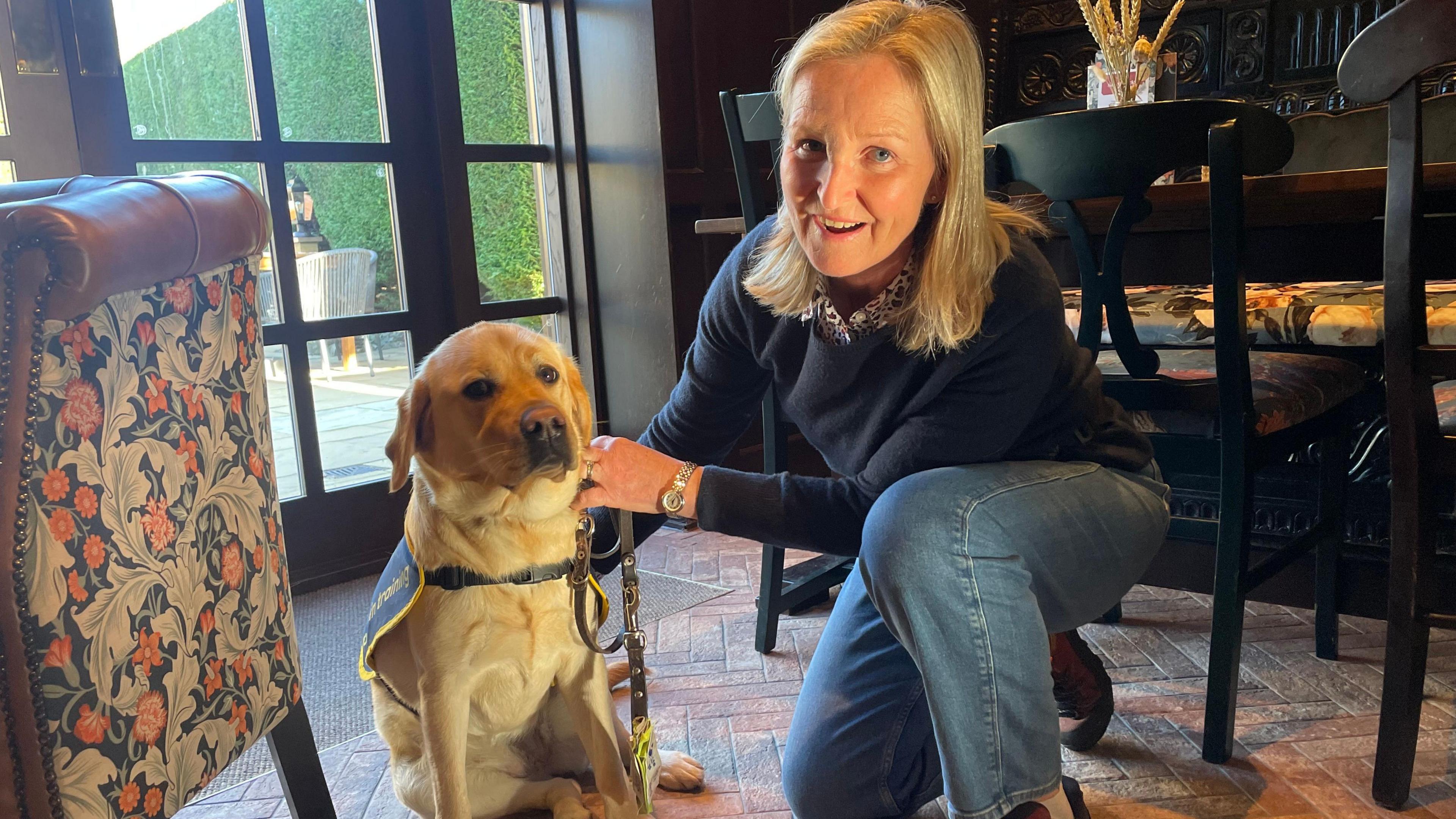 A woman kneels on one knee next to her Labrador puppy. She is wearing a navy blue jumper and blue jeans and is smiling.