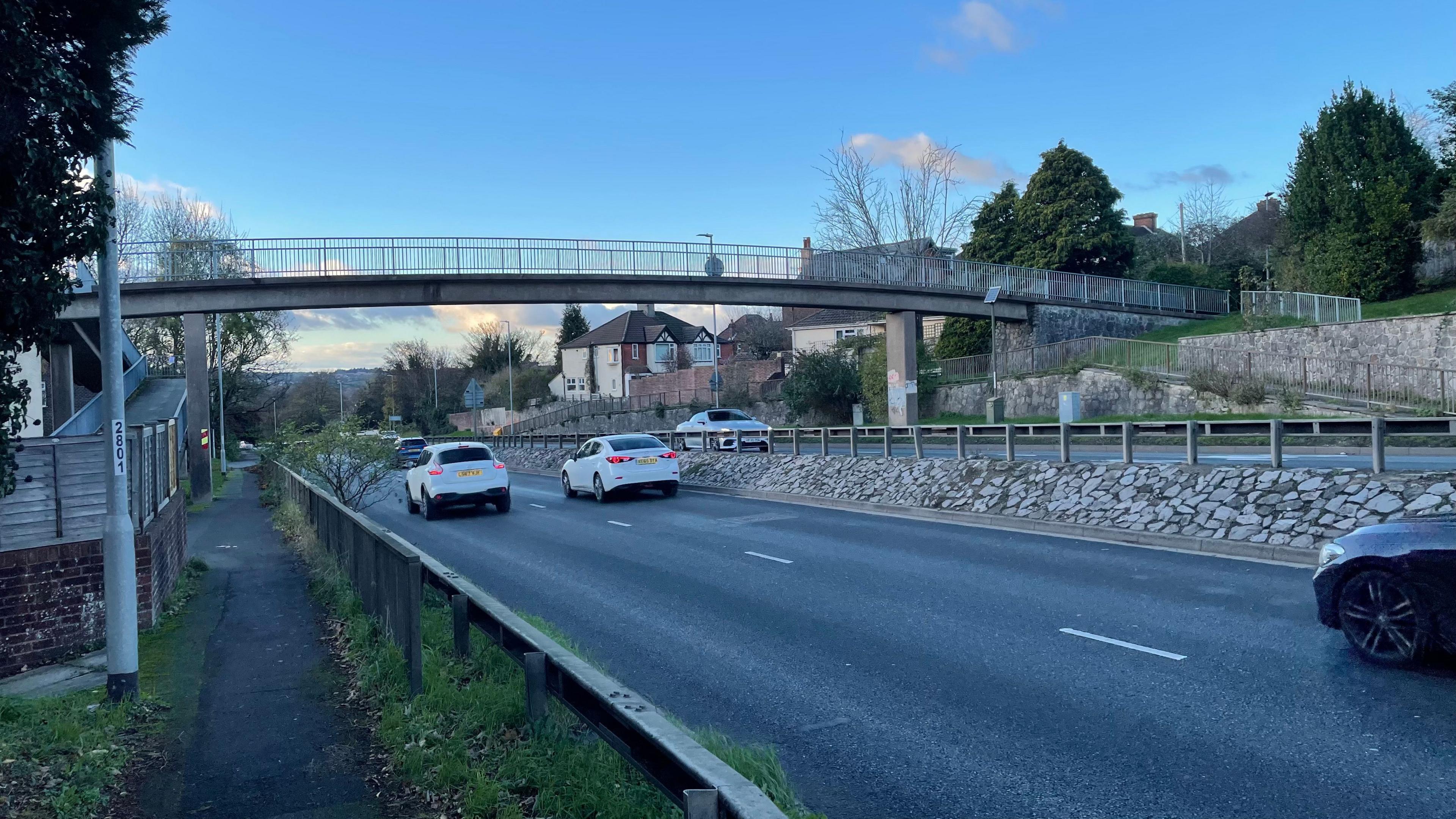 A two lane road with cars driving away from the camera, with a grey footbridge above the road and a blue sky with clouds in the background.