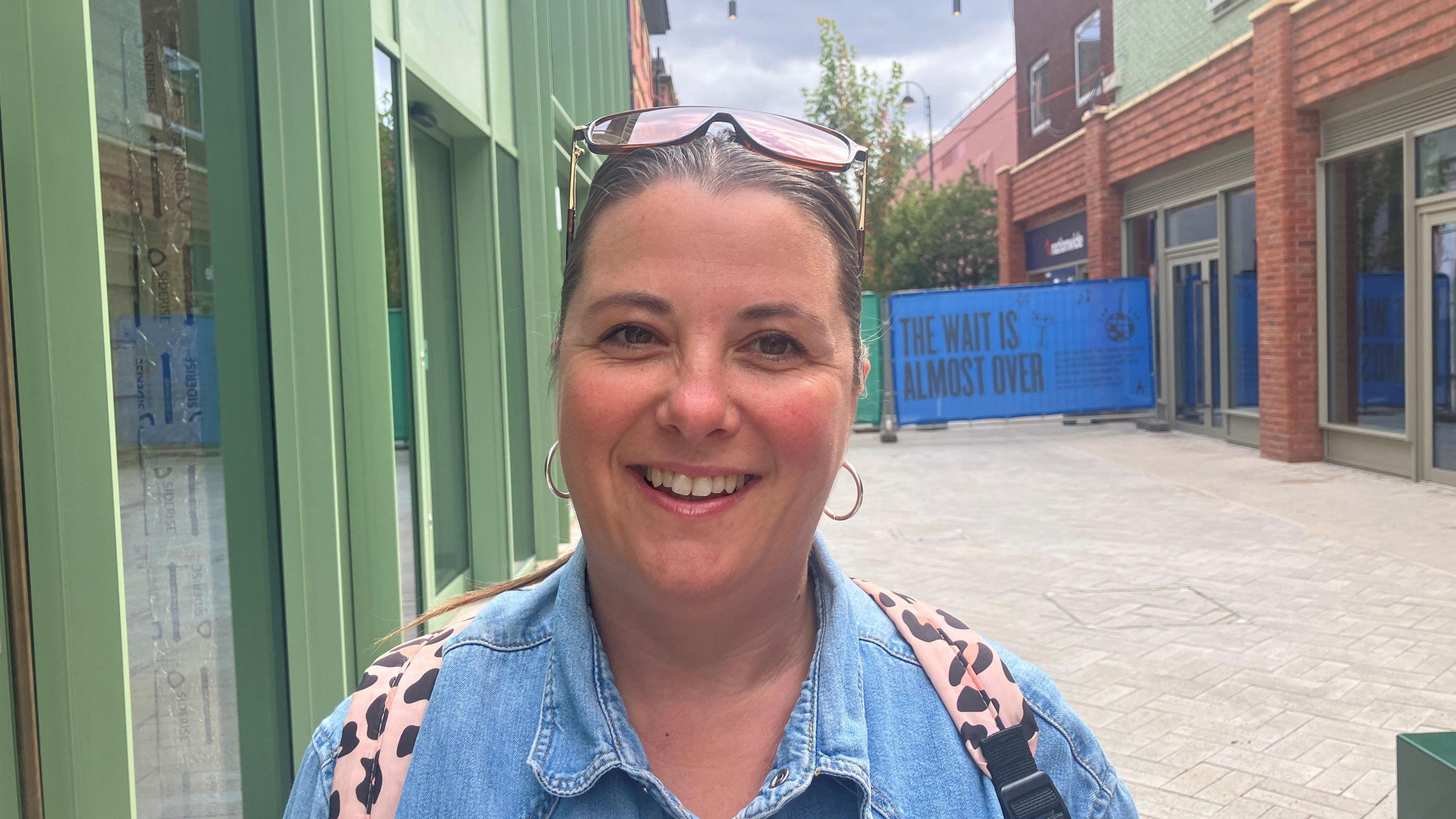 A woman wearing a blue denim top and a backpack stands smiling on a street.