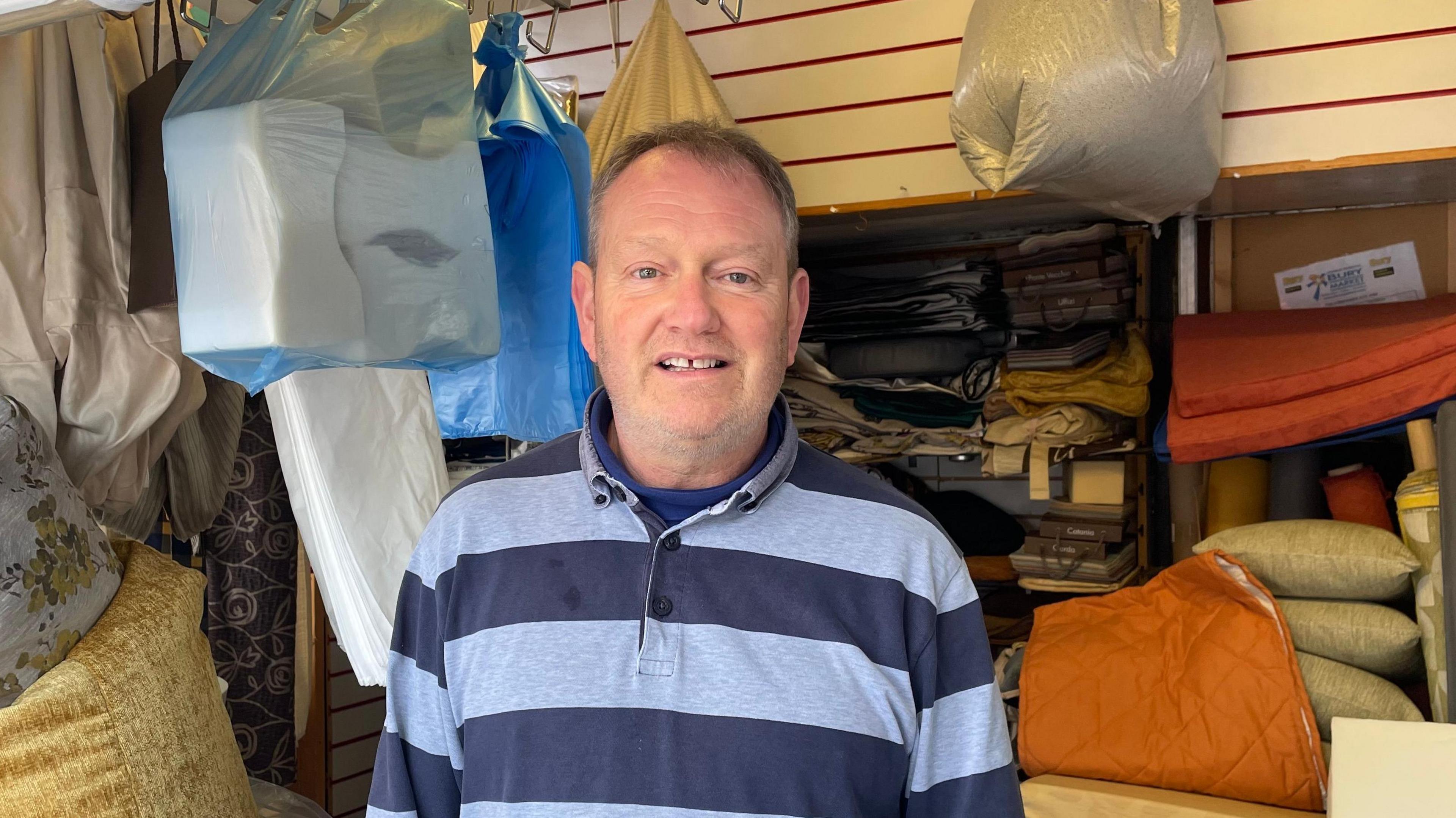 Steve Taylor wearing a striped blue and grey polo shirt stood in a room in his shop surrounded by cushions and bags of foam material.