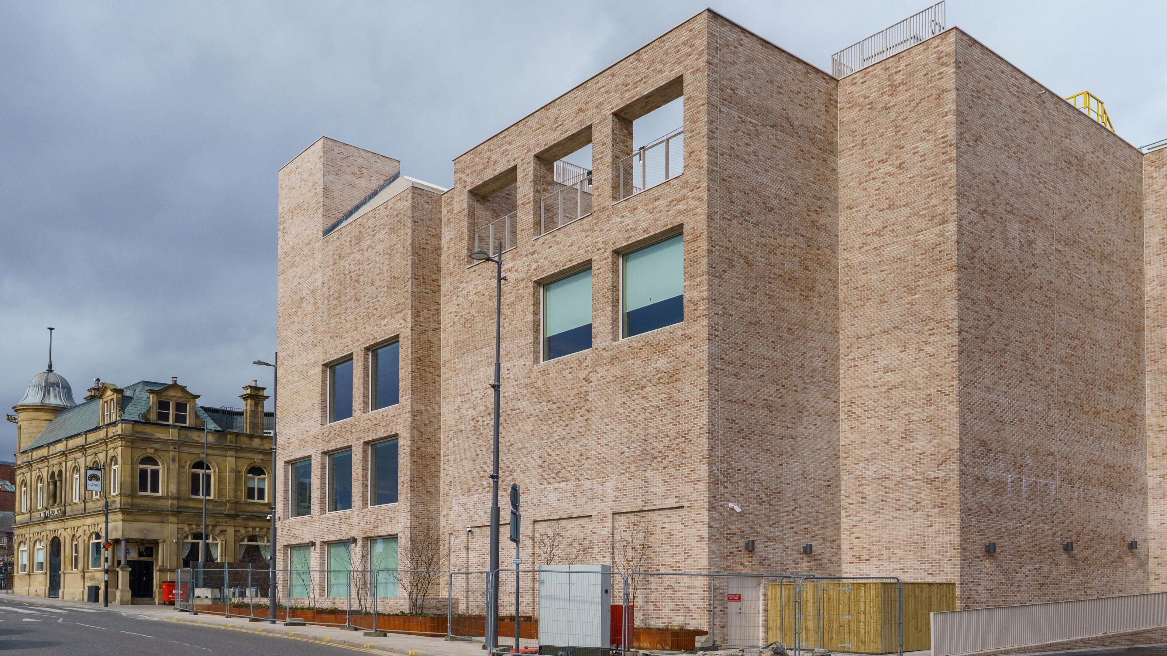 The multi-storey Culture House photographed from South Street behind Keel Square. The brick building is fenced off. The sky is grey. It has large square windows. There is another small building next to it. It has more decorations and looks less modern.