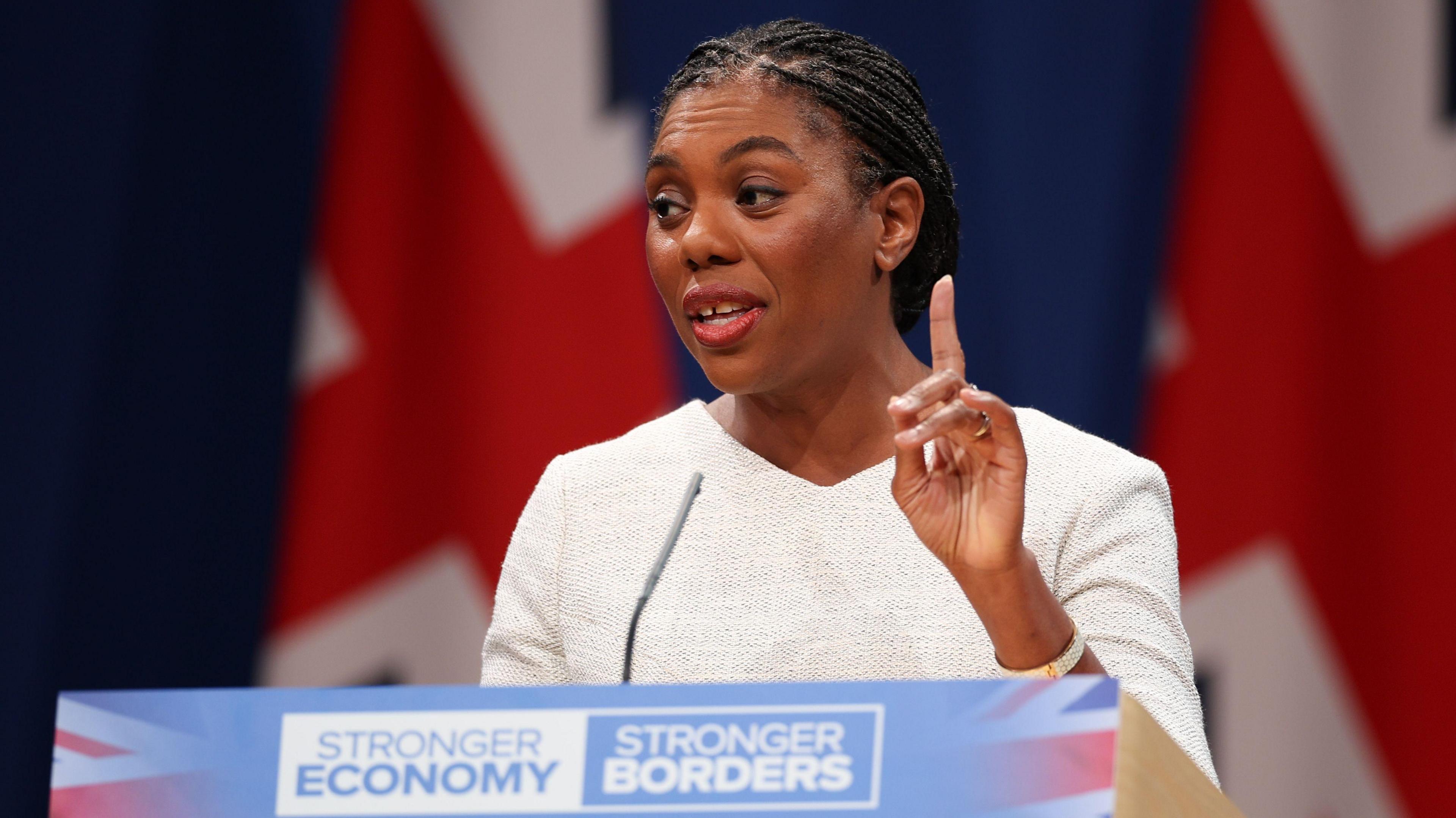 Kemi Badenoch stands at a podium with two British flags behind her and raises one finger in front of her. She wears a white shirt. The podium reads "stronger economy" and "stronger borders".