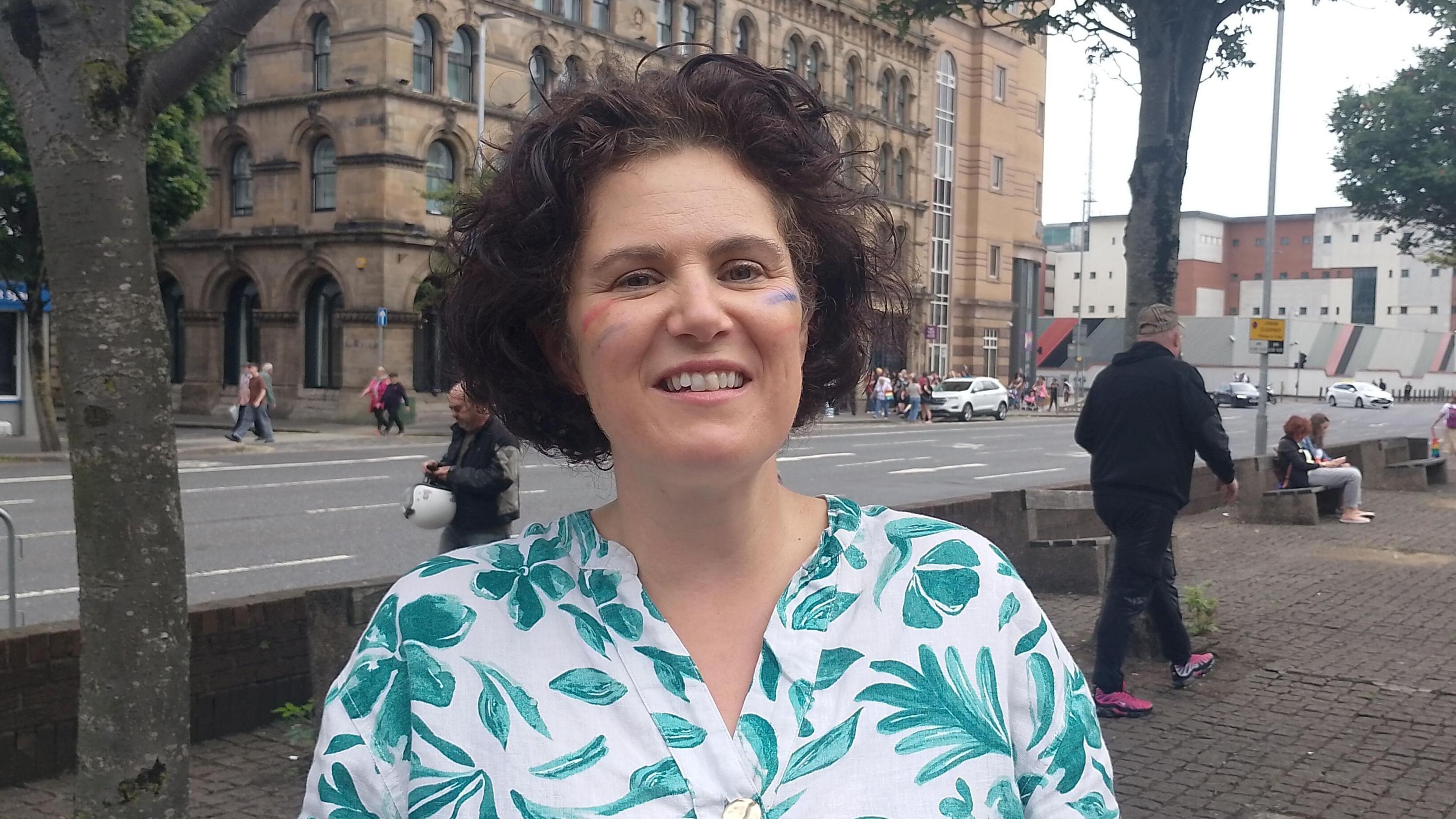 Claire Hanna. She has short brown curly hair, wearing a green floral blouse. She has a rainbow on her cheek.