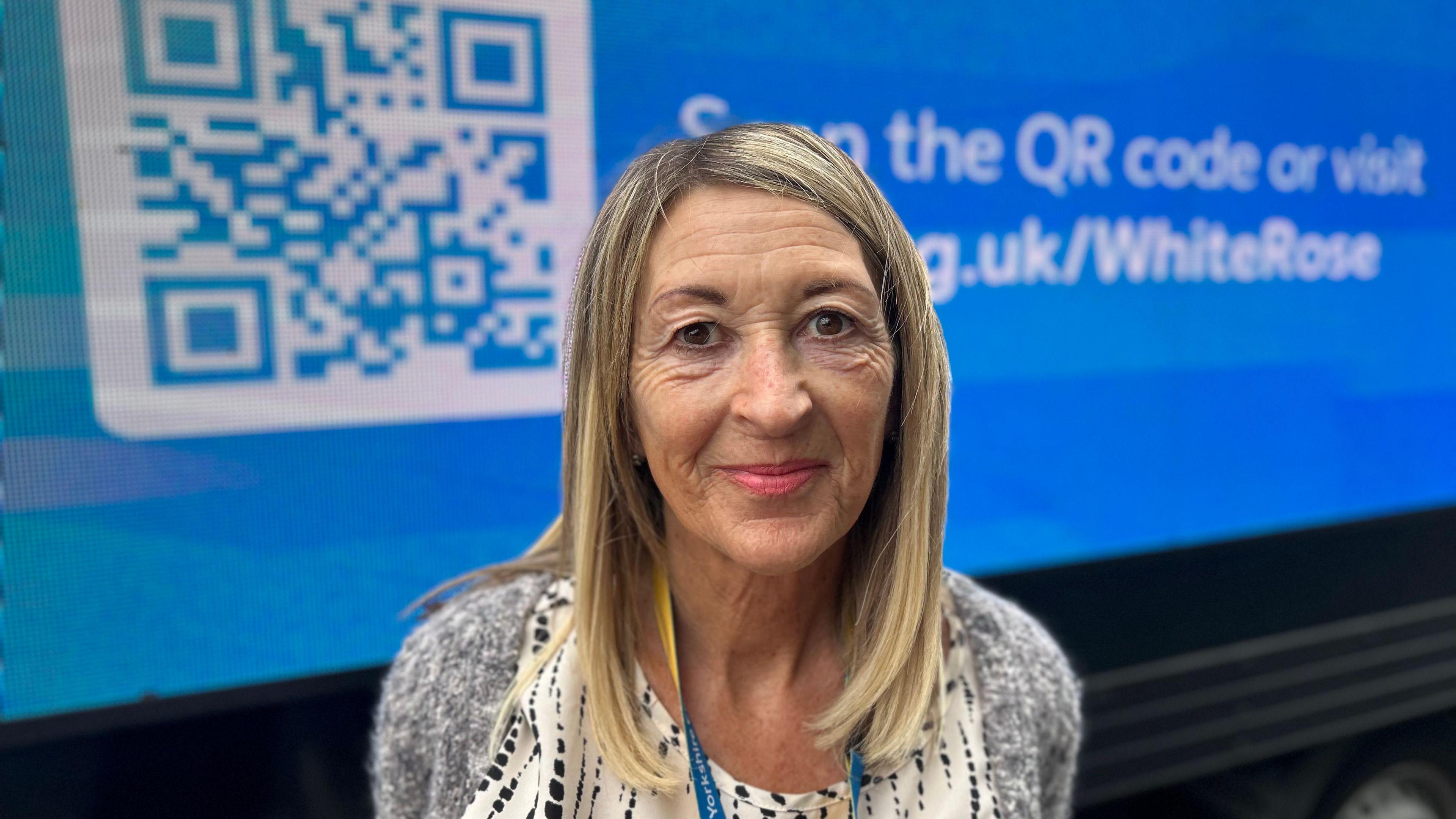 A woman with long blond hair and wearing a grey jumpers smiles as she stands in front of a blue van. On the side of the van is displayed a large white QR code  and text (partially obscured) reading, "Scan the QR code or visit ycr.org.uk/whiterose".