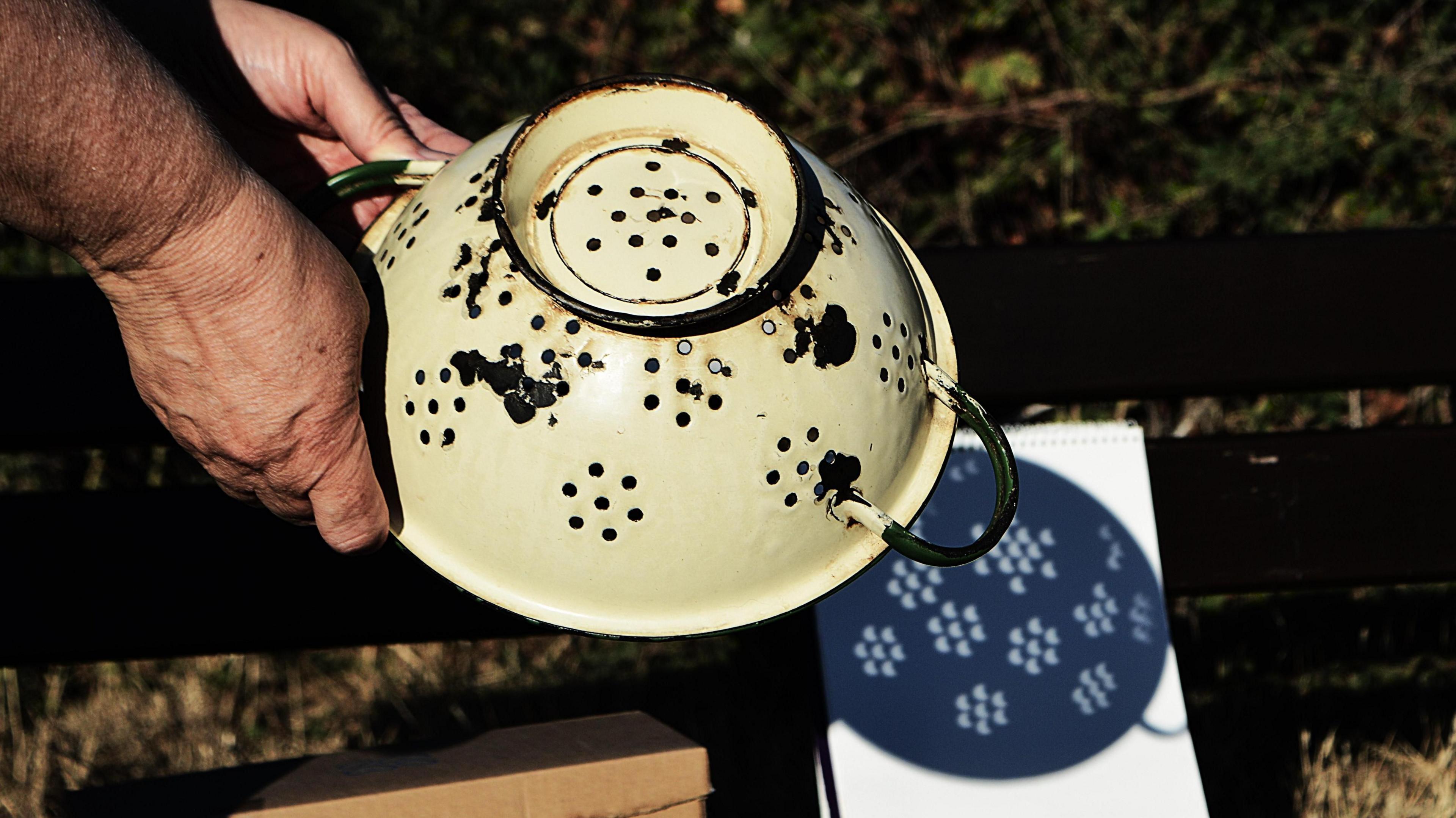 A person holds an upside-down, cream-coloured metal colander above a sheet of white paper in Hastings, East Sussex. Sunlight passes through the holes in the colander, creating dozens of crescent-shaped projections that show the partially eclipsed Sun.