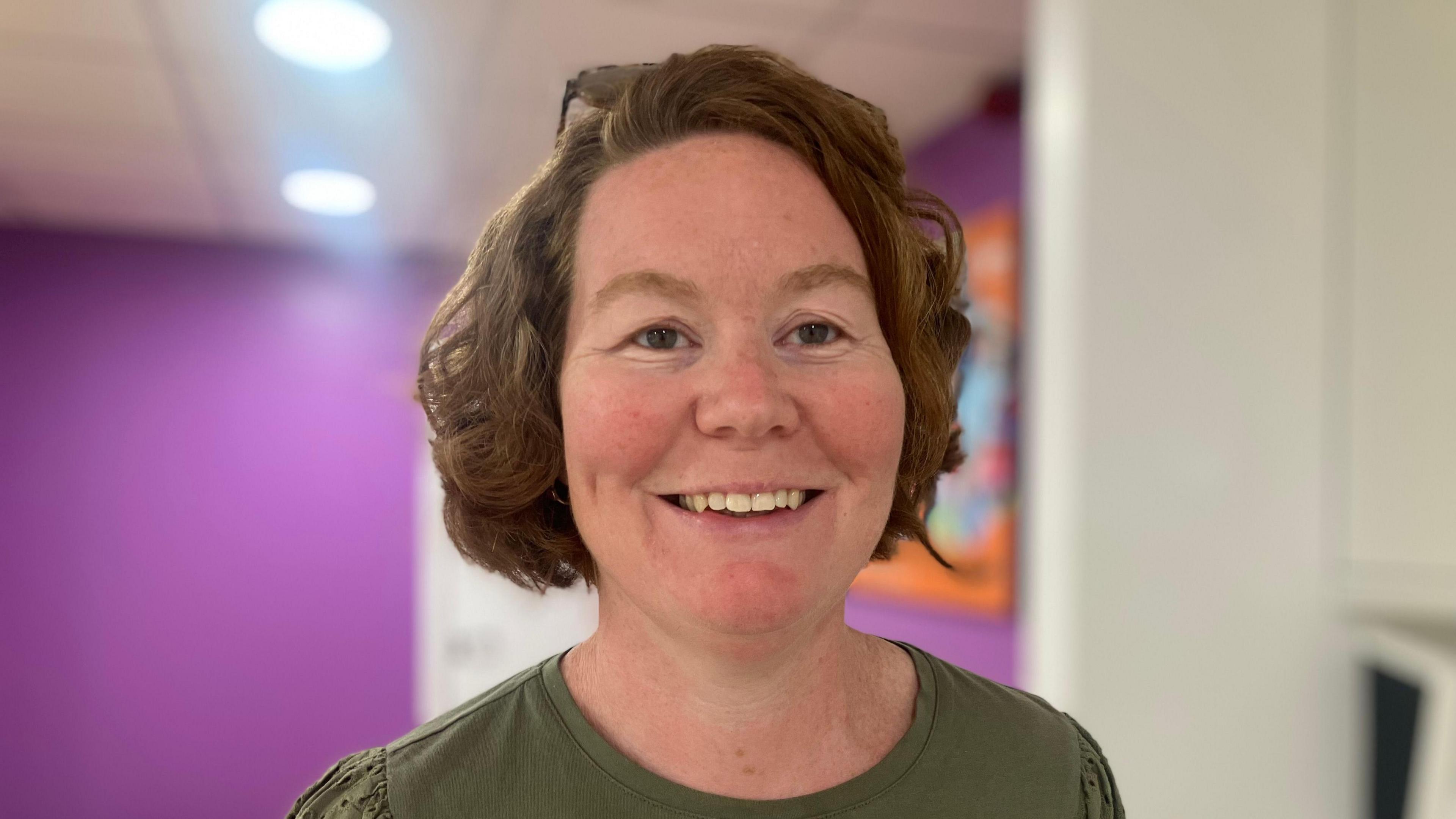 A woman with red hair smiles at the camera. Behind her, a purple wall is illuminated by overhead lights. She wears a green top.