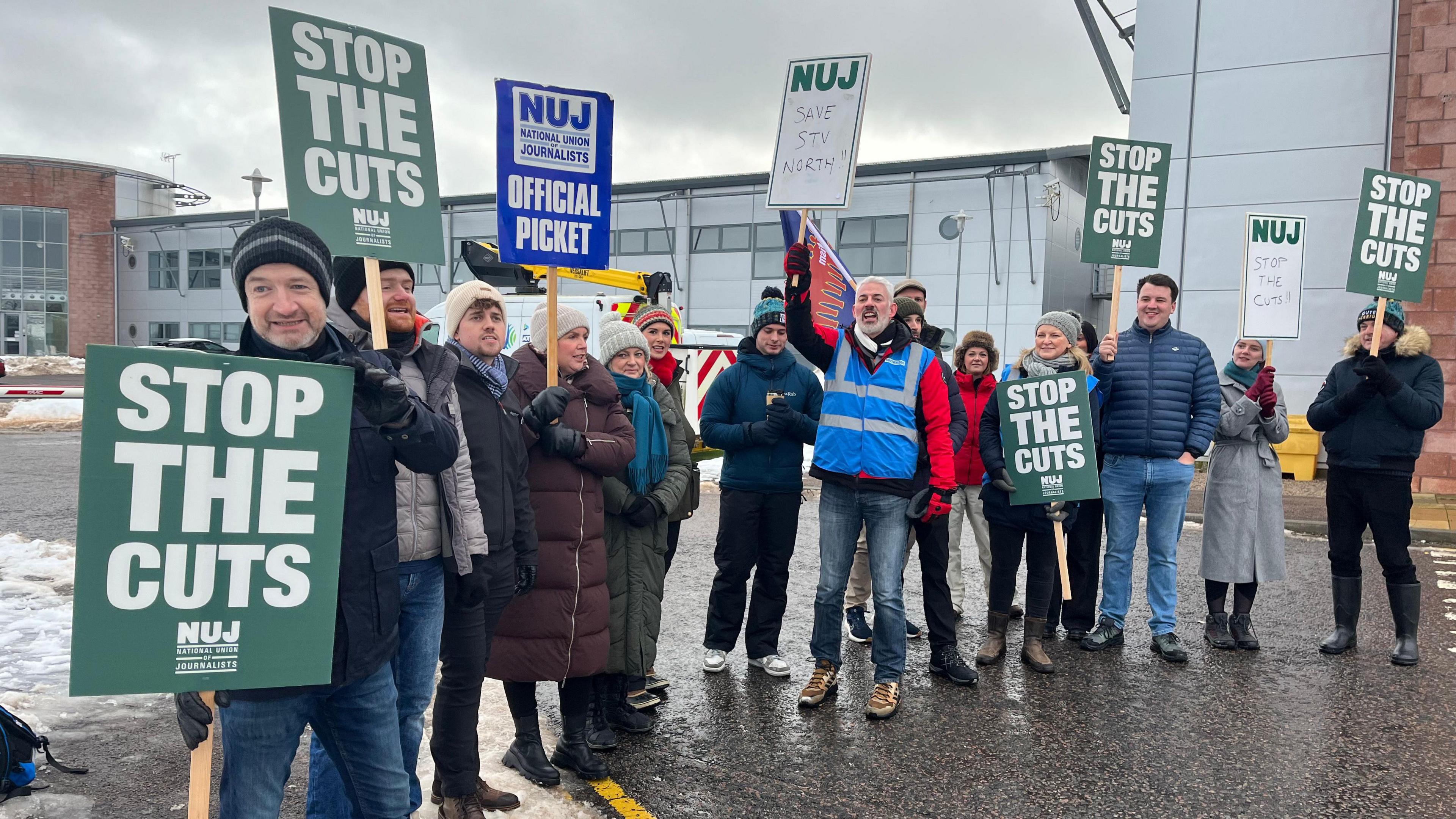 A group of picketers stand in a row holding strike placards. They are in front of a modern building and there is snow on the ground.