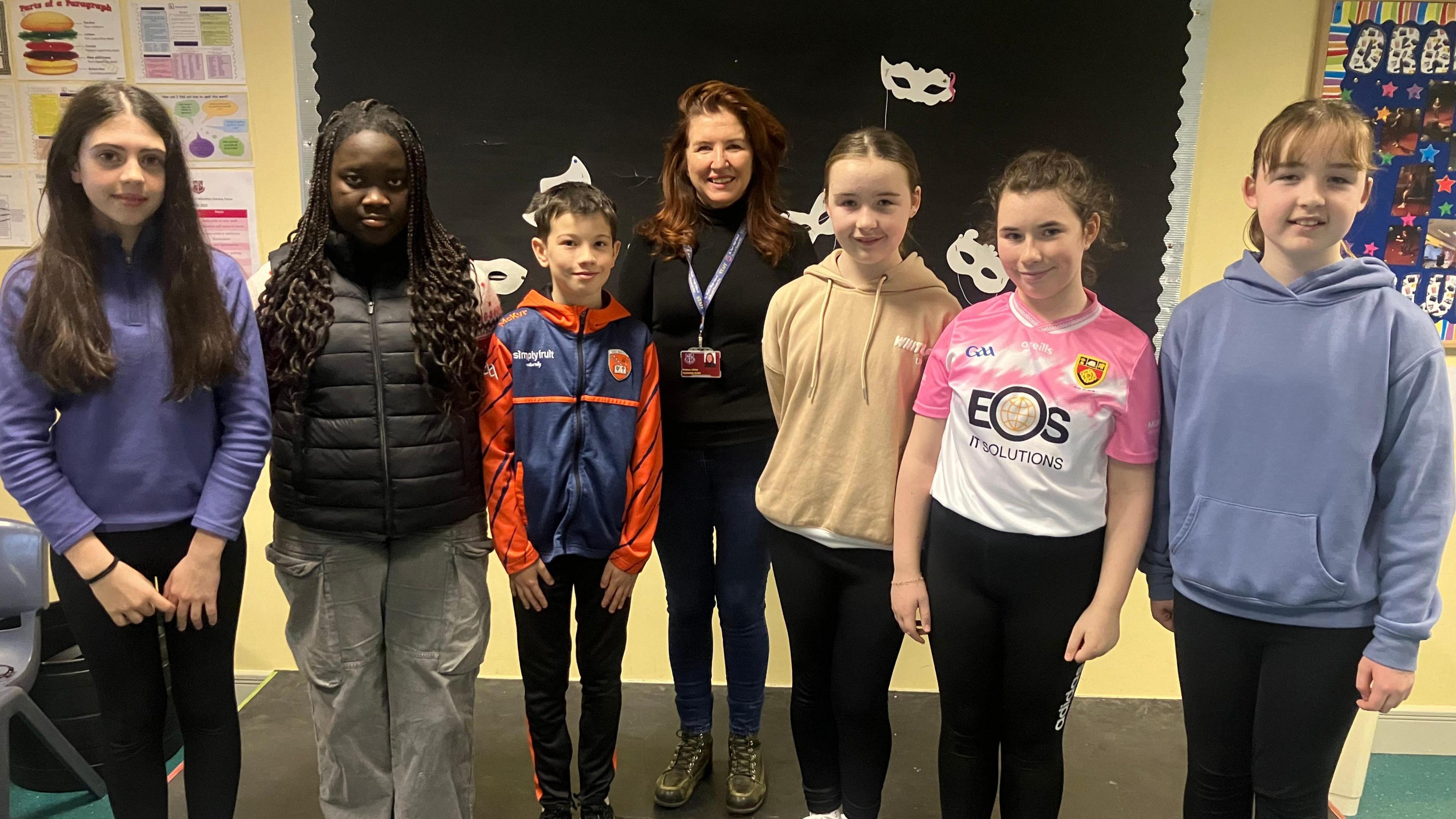 School children standing next to each other with their teacher in the middle. To the left of the teacher is three female pupils with tied back hair wearing hoodies and one wearing a sports top. On the right of the teacher, who has long ginger hair and is wearing black, is two female students and a male, all are wearing a sip-up top and a body warmer.