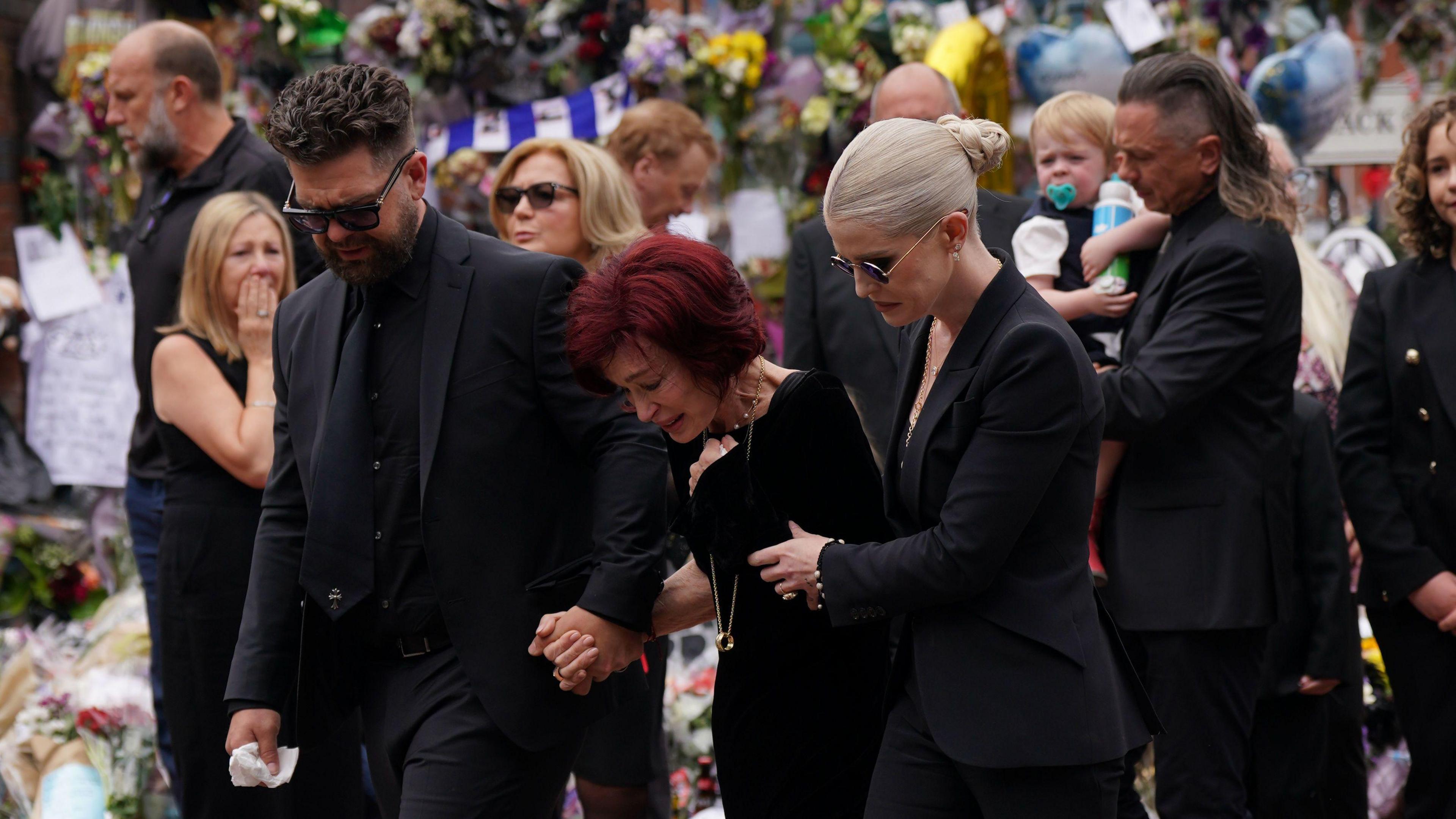 Sharon Osbourne who has red hair, is wearing black and bent over crying. On one side of her is her son who is in all-black, with black sunglasses, on the other side is her daughter Kelly who has white blonde hair in a bun. She is also wearing all black. In the background are dozens of people crying, as well as floral tributes and gifts