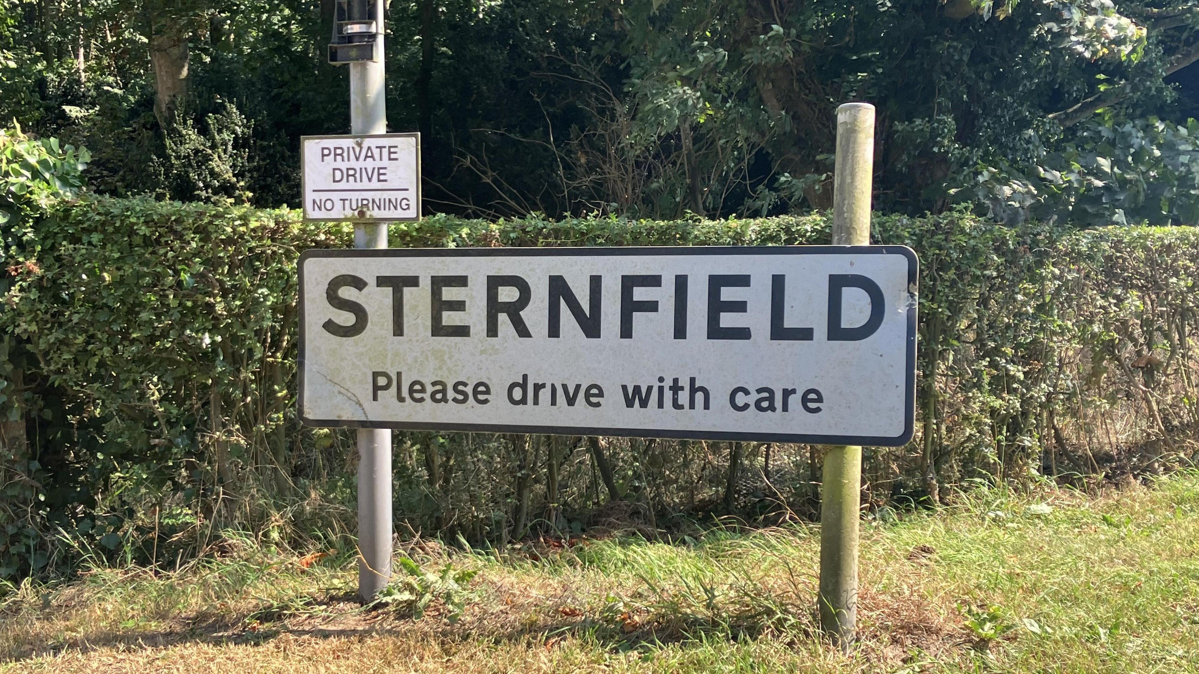 Village sign reading "Sternfield Please Drive with Care" set on a grass verge in front of a hedge