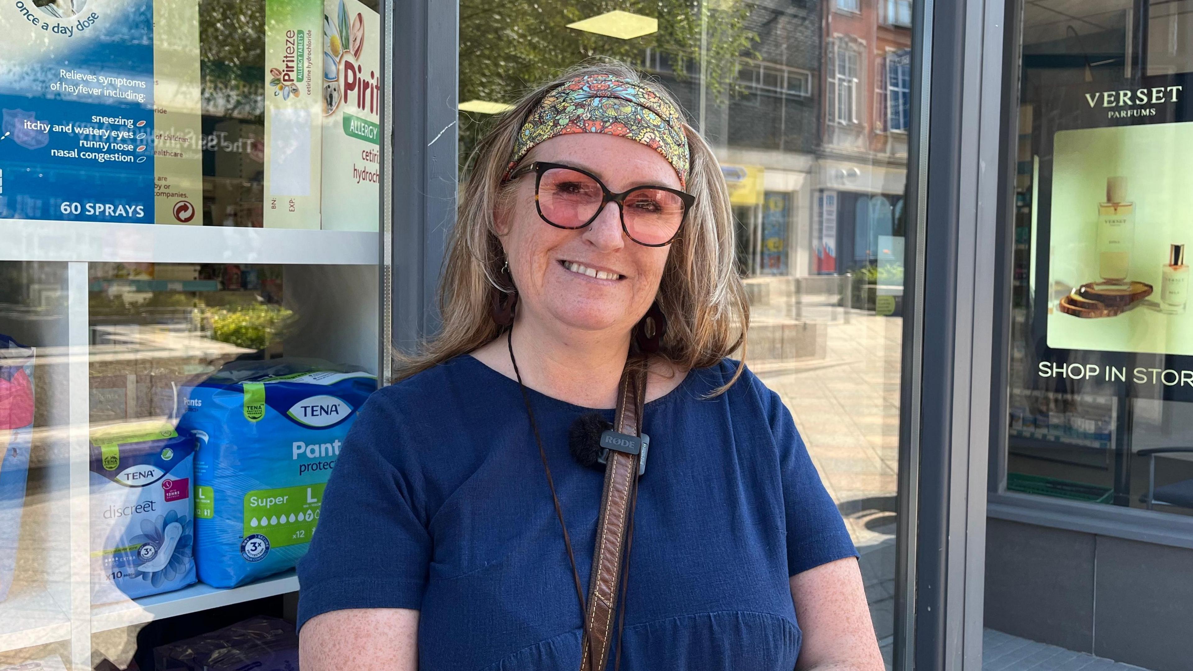 A person stands outside a pharmacy, wearing glasses and a patterned headband, with healthcare products displayed in the shop window behind them.
