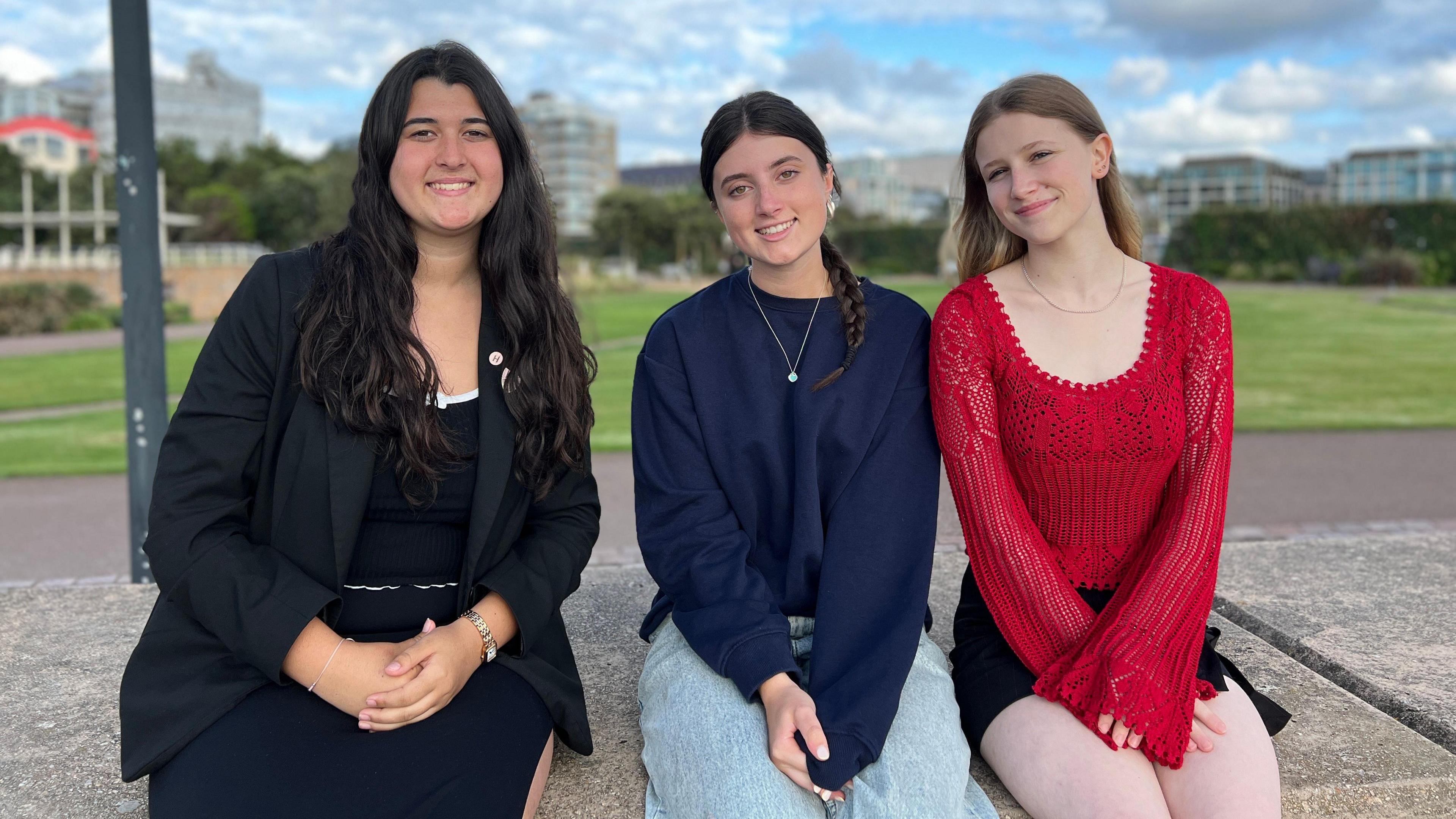 The three girls sit on a concrete wall and smile at the camera. Saskia is on the left and is where a black dress, top and jacket. Tilly is in the centre and has baggy jeans and a baggy blue jumper and wears a necklace. Lily is on the left and is wearing a red laced top and has black shorts on. Behind them is a green patch of grass with buildings in the background.