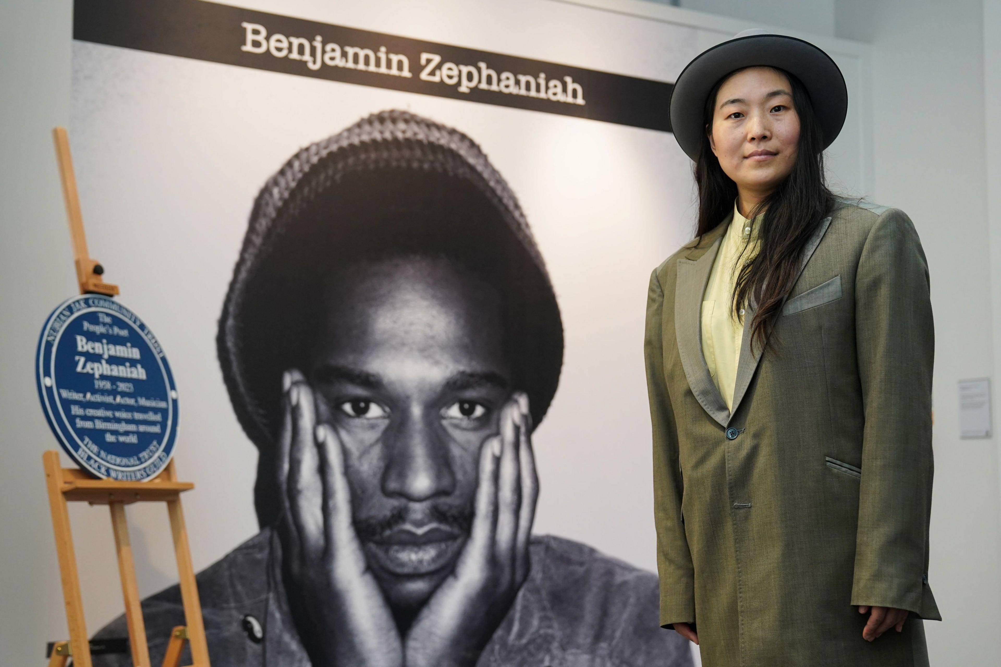 Qian Zephaniah wearing a brimmed hat and green-grey jacket in front of a portrait of Benjamin Zephaniah and blue memorial plaque at a recent event.