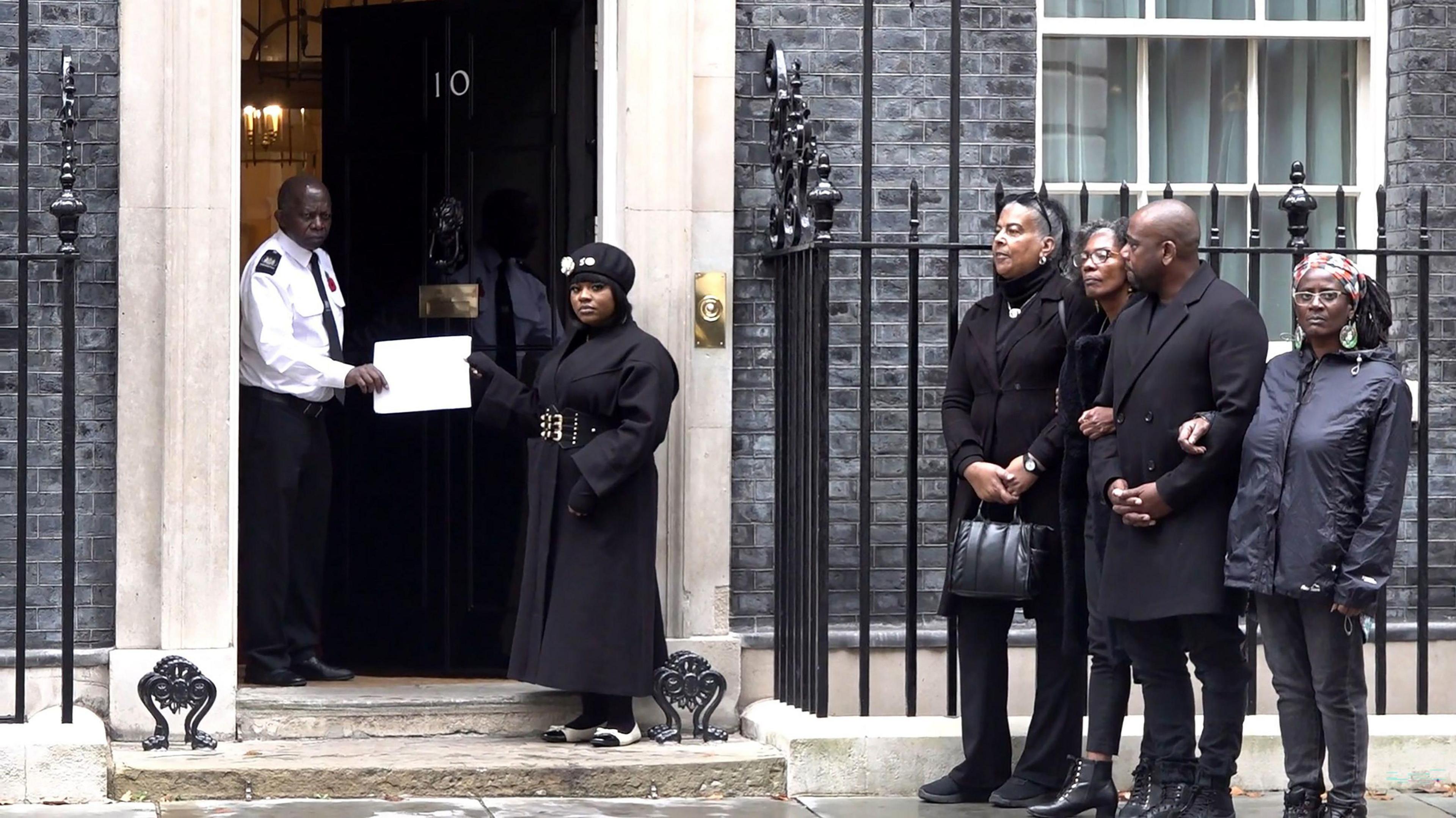 Four people stand together outside 10 Downing Street while a woman dressed in black hands a large white envelope to a security guard in the doorway