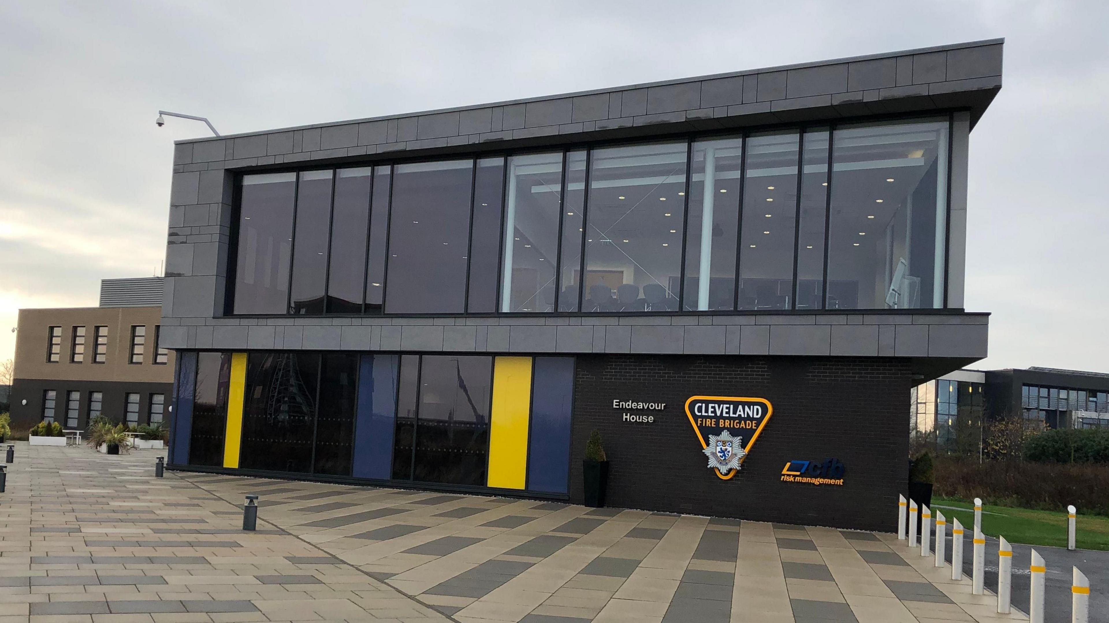 Cleveland Fire Brigade Headquarters. A large black cladded building with wide windows on the top floor. A sign on a wall next to a large windowed entrance door reads 'Cleveland Fire Brigade'.