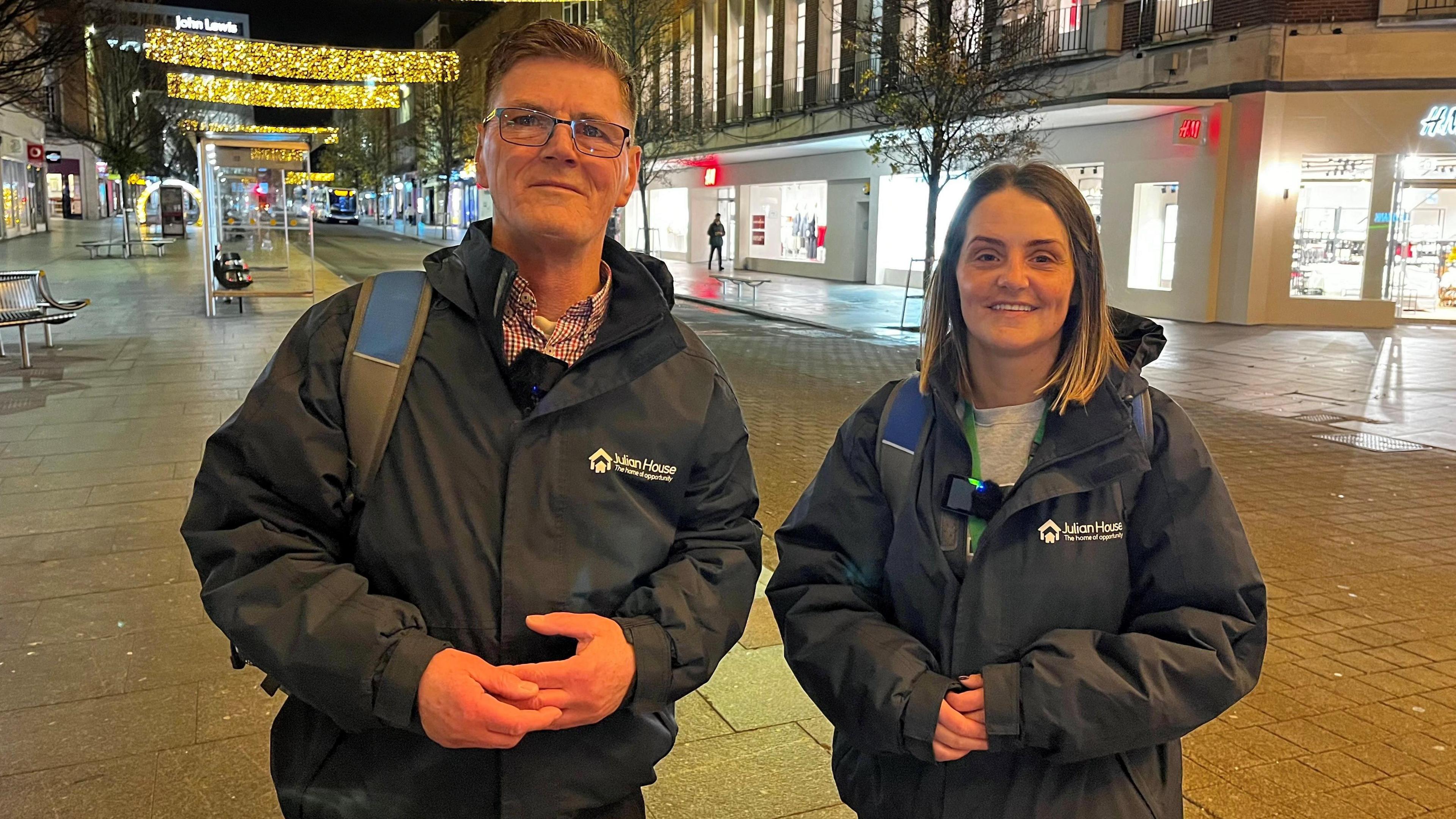 Jim McCarthy (left) and Sophie Wisdom (right) stand on Exeter High Street early in the morning. They are stood in front of an H&M store and are wearing Julian House branded navy coats. Christmas decorations can be seen in the background along the High Street along with a bus stop. Jim is wearing glasses.