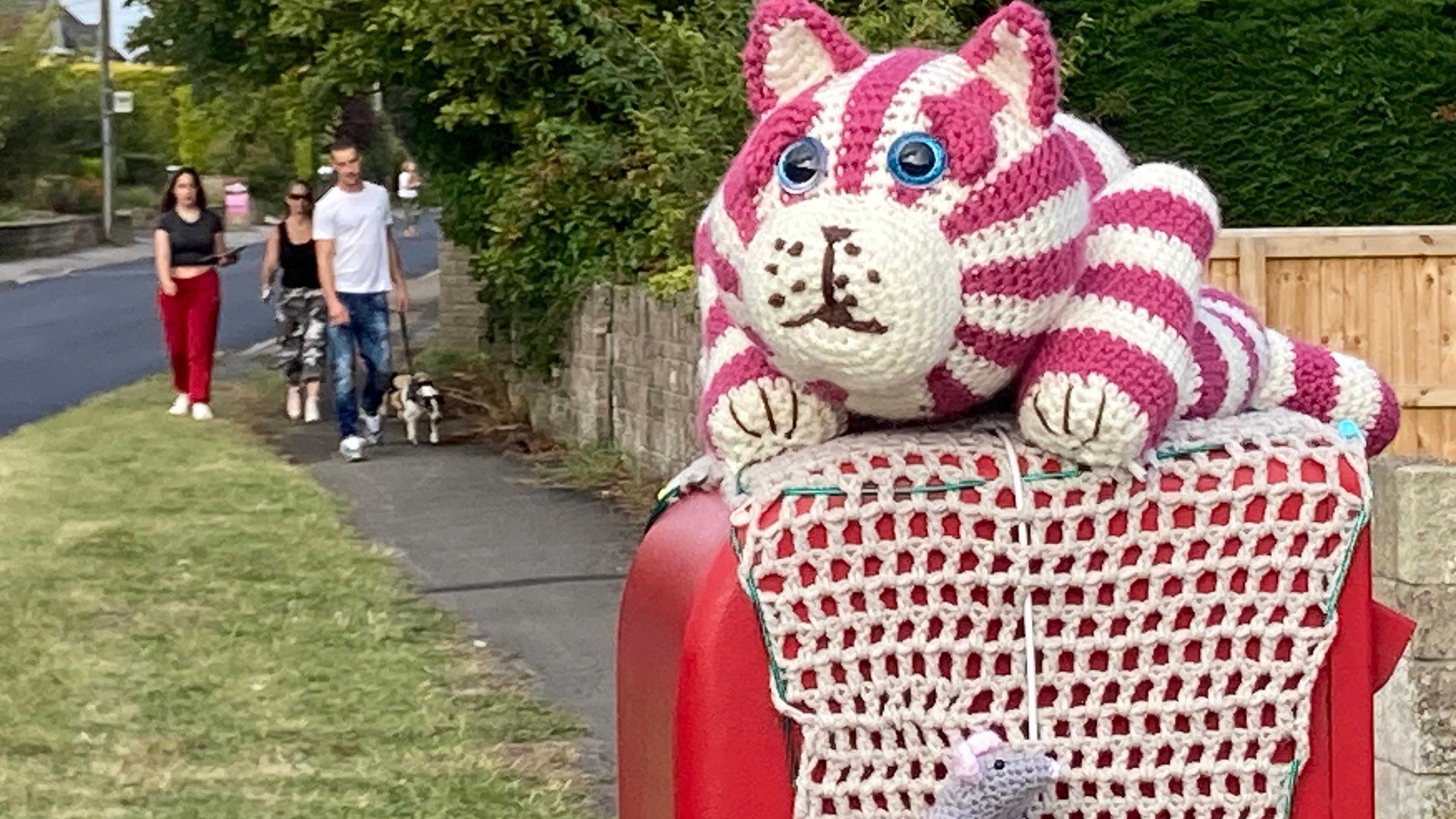 A knitted Bagpuss with a mouse on top of a postbox