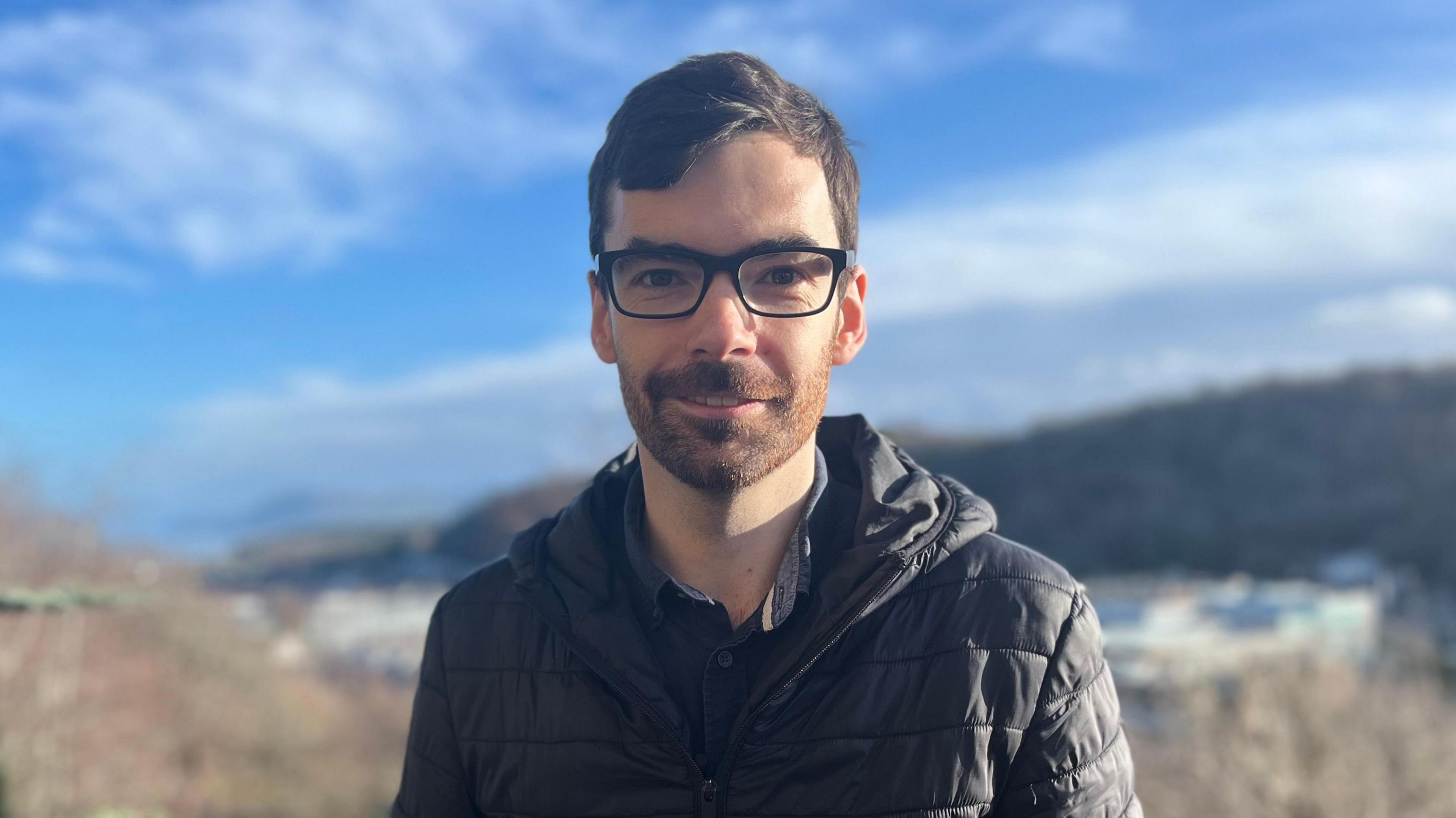 Dyn yn gwisgo cot ddu gyda barf a sbectol. Mae'n sefyll tu allan gydag awyr las a bryniau yn y cefndir