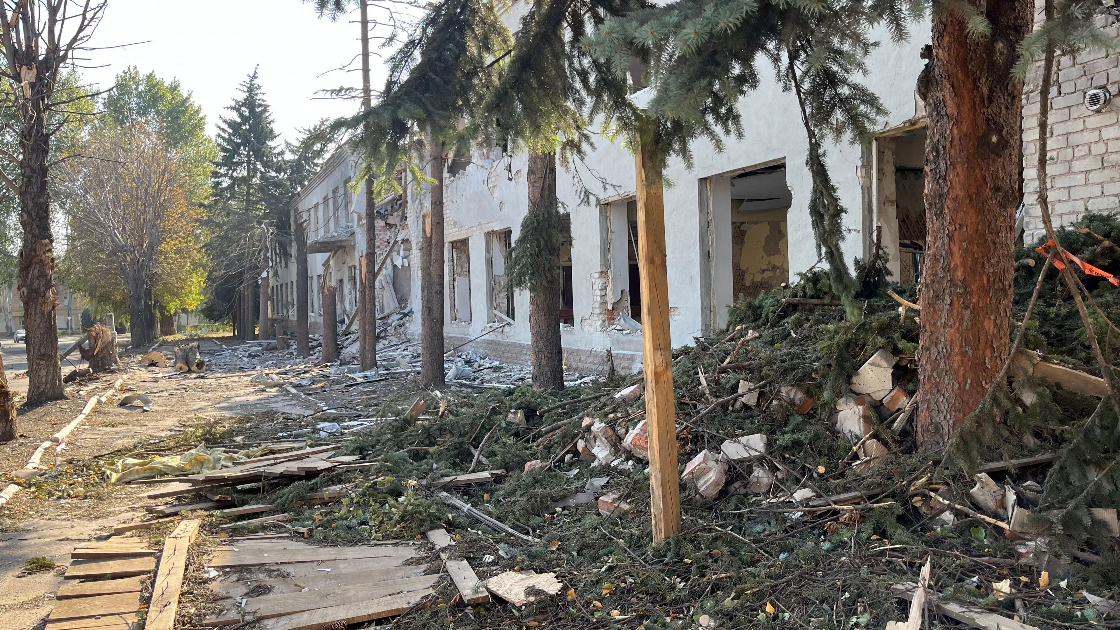 Daylight: an empty street in Pokrovsk filled with piles of rubble and broken branches from damaged buildings and trees. Windows have no glass.