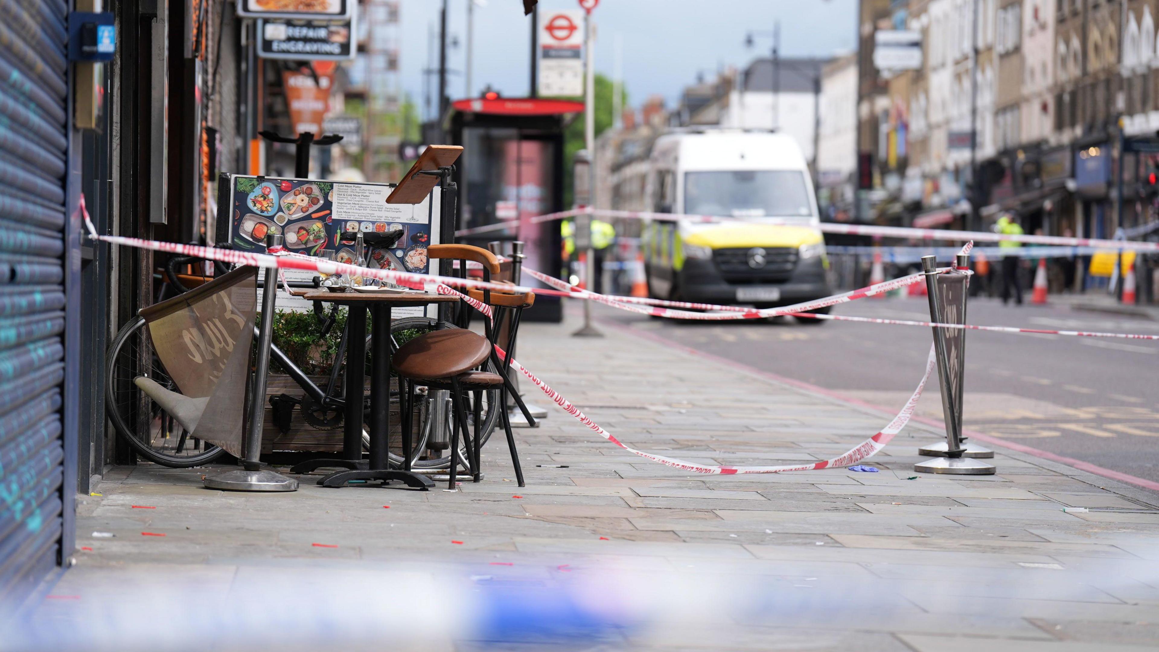 An exterior view of the restaurant where the victims were attacked in Dalston. There is a police tape cordon in place, and a police van blurred in the distance, There is debris on the pavement.