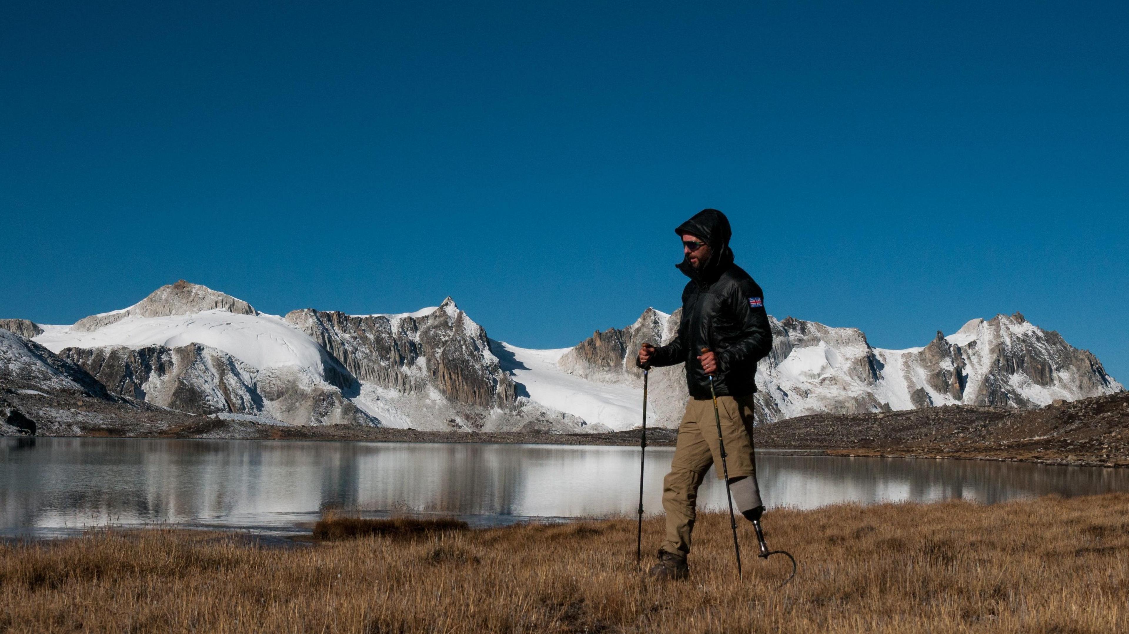 Rich Potter, who is wearing a black-hooded coat, sunglasses, and light brown trousers, walking in front of snow-covered hills and a lake. He is wearing a black boot on his left foot and has an artificial right leg. 