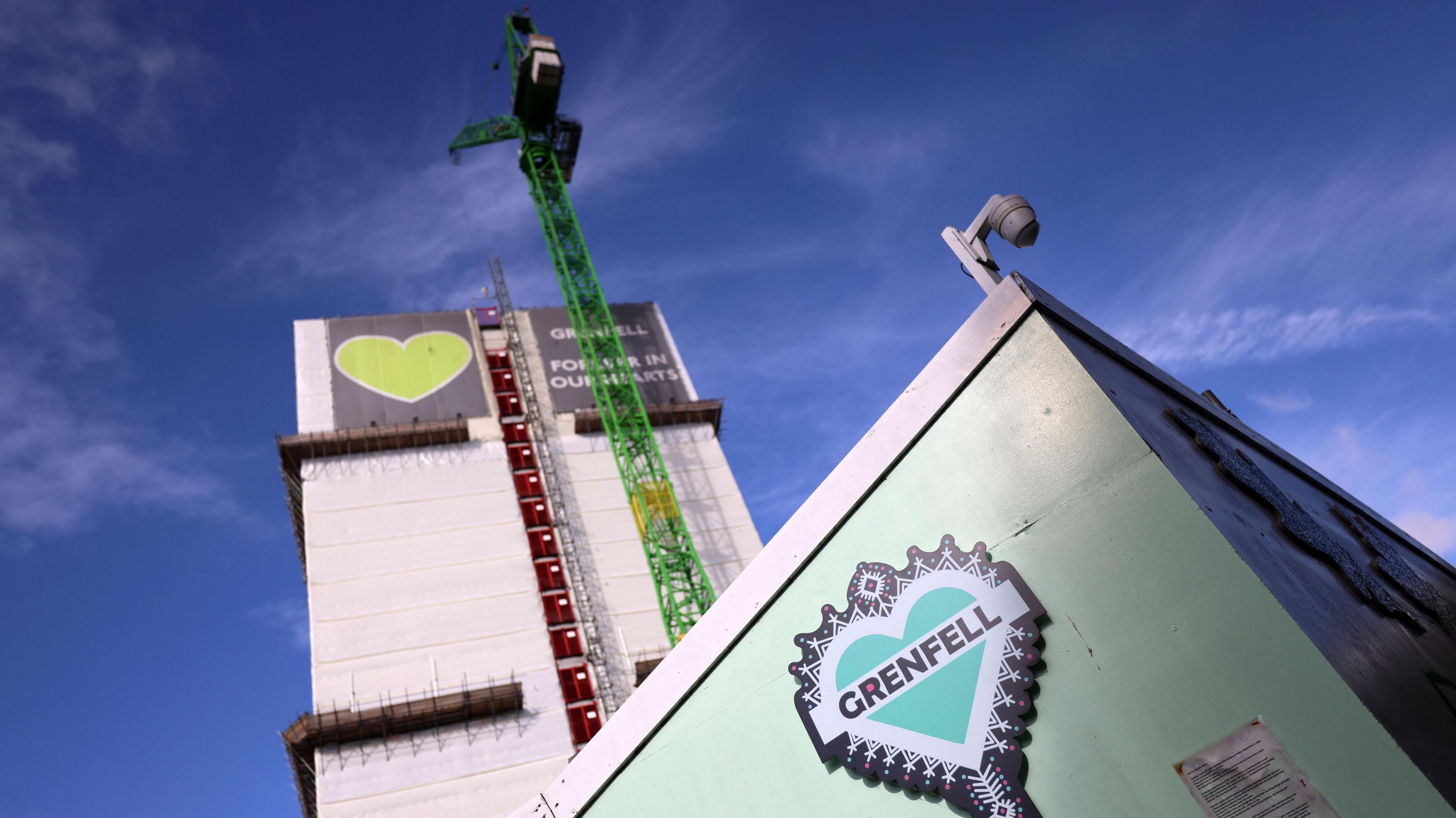 A green crane runs alongside Grenfell Tower with a large, green heart visible on the top of the building and on a hoarding protecting the building at street level. Blue sky can be seen behind it
