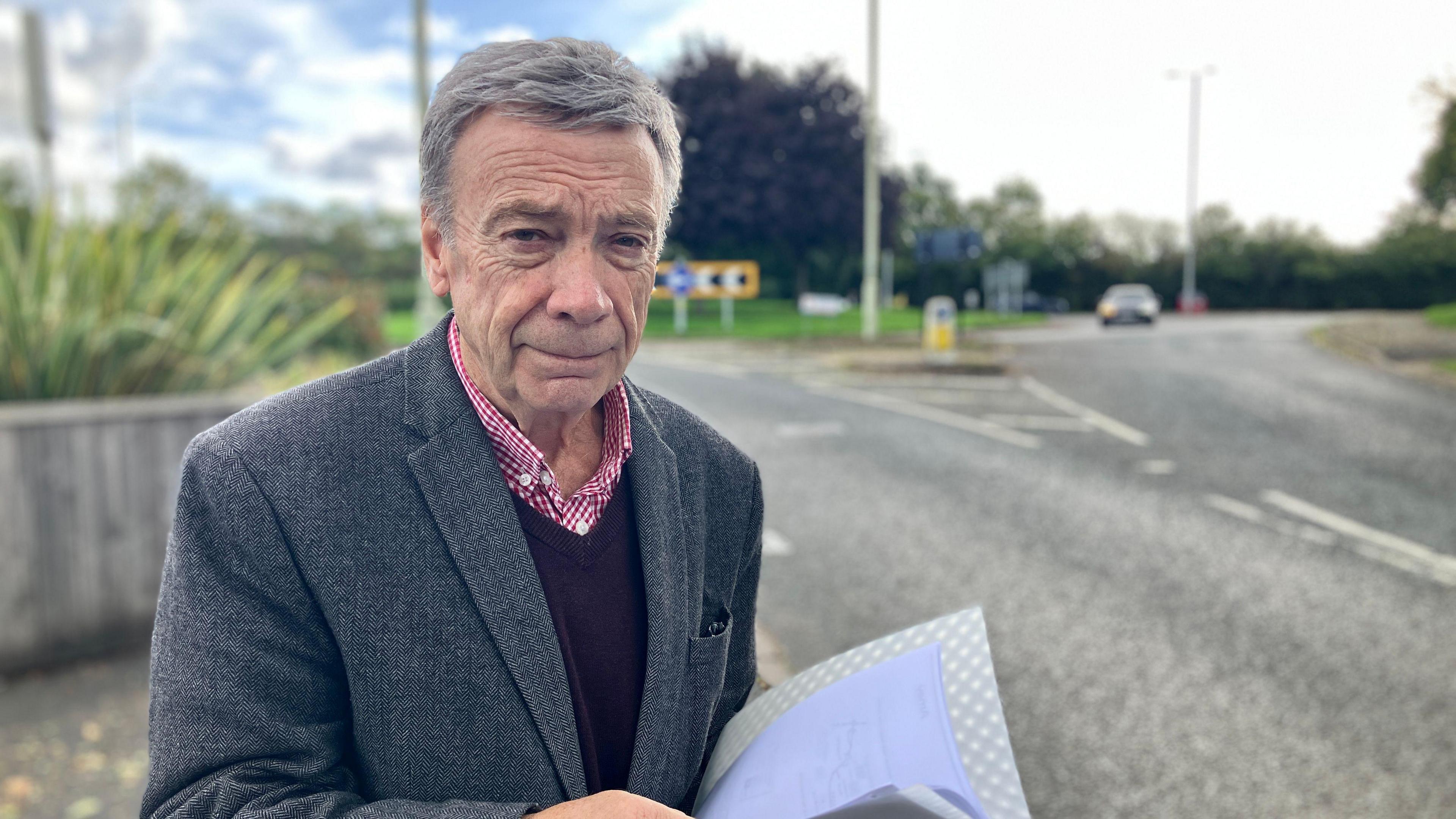A grey-haired man, wearing a dark grey blazer, black v-neck jumper and red shirt, whilst holding a notepad, is standing in front of a roundabout with lampposts and a bollard